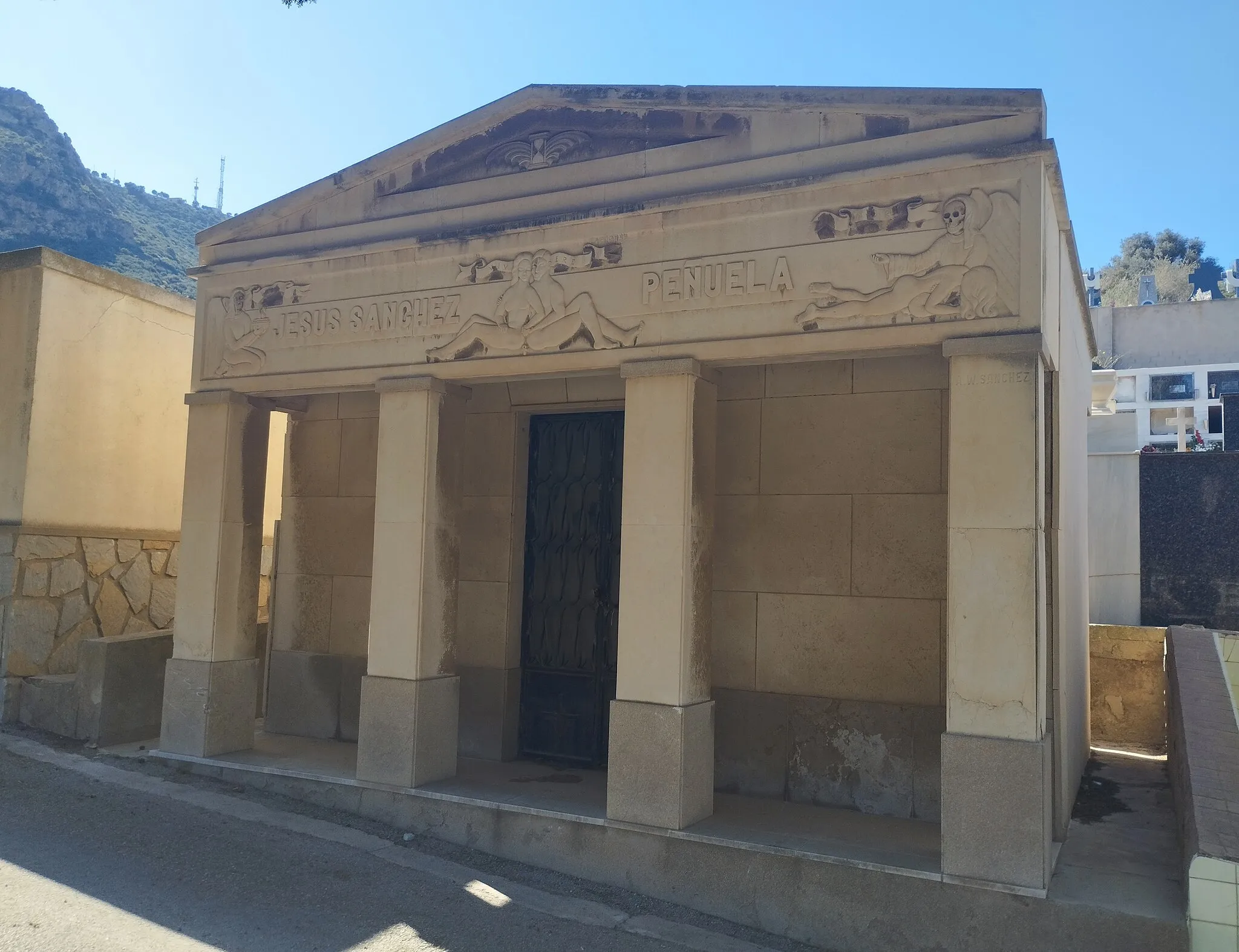 Photo showing: The Mausoleum of the pharmacist Jesús Sánchez Peñuela, in the Los Remedios Cemetery in Cartagena (Spain), is a building that was built in the 1940s or 1950s with the participation of the sculptor Agustín Wenceslao Sánchez Velázquez and an unknown architect. Its tetrastyle facade stands out, with four square columns that support an entablature decorated with allegorical reliefs of life and death, while a winged hourglass is represented on the pediment.