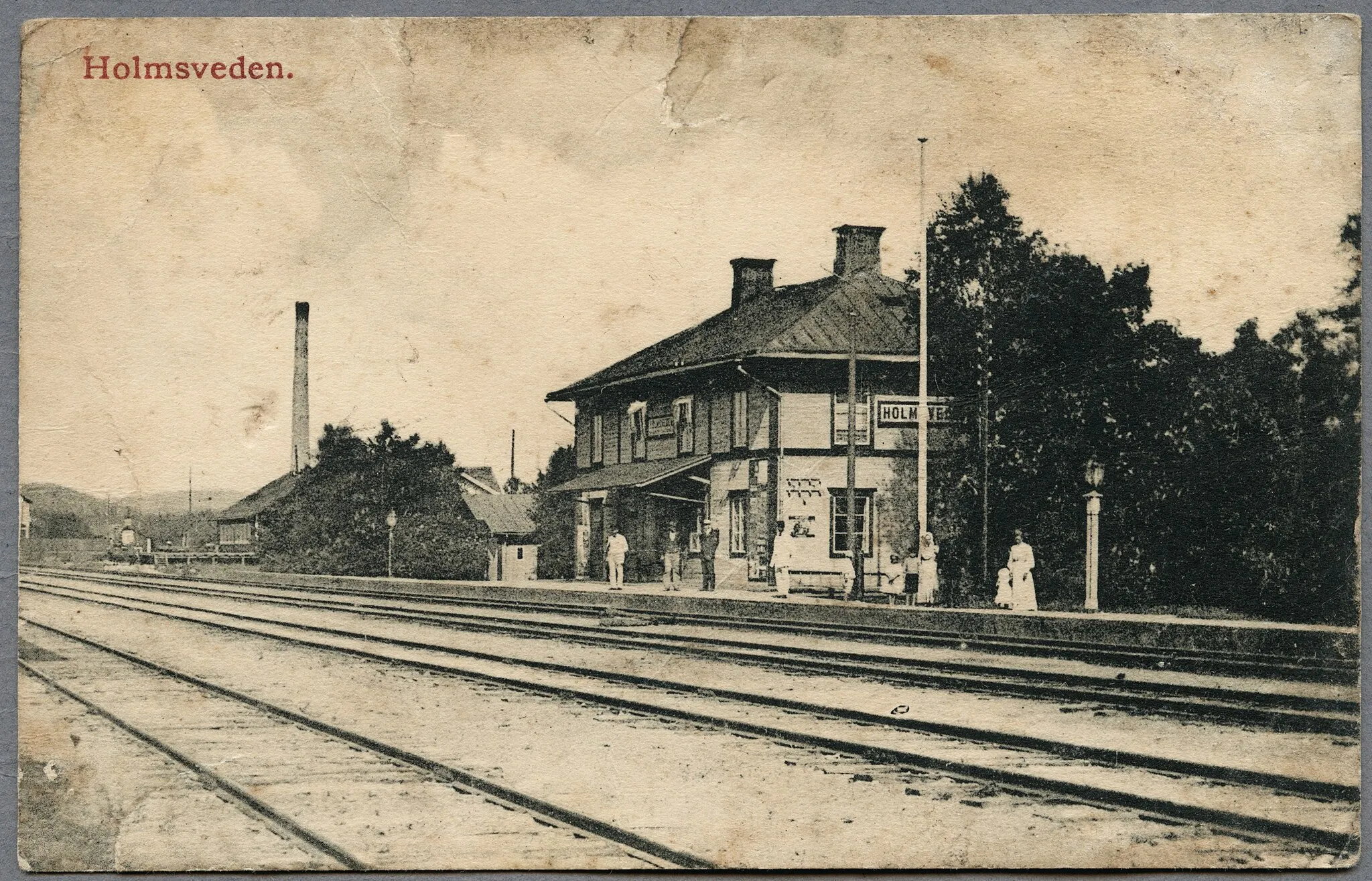 Photo showing: Det svartvita fotografiet visar järnvägsstationen i Holmsveden en sommar omkring 1910. Väntande på perrongen framför stationsbyggnaden står tio personer varav fyra barn. I bakgrunden sticker en hög murad industriskorstenen upp. Fotografen står på den sydvästra sidan av järnvägsspåren och avbildar stationsbyggnaden, på den nordöstra sidan, snett över spåren i nordlig riktning.