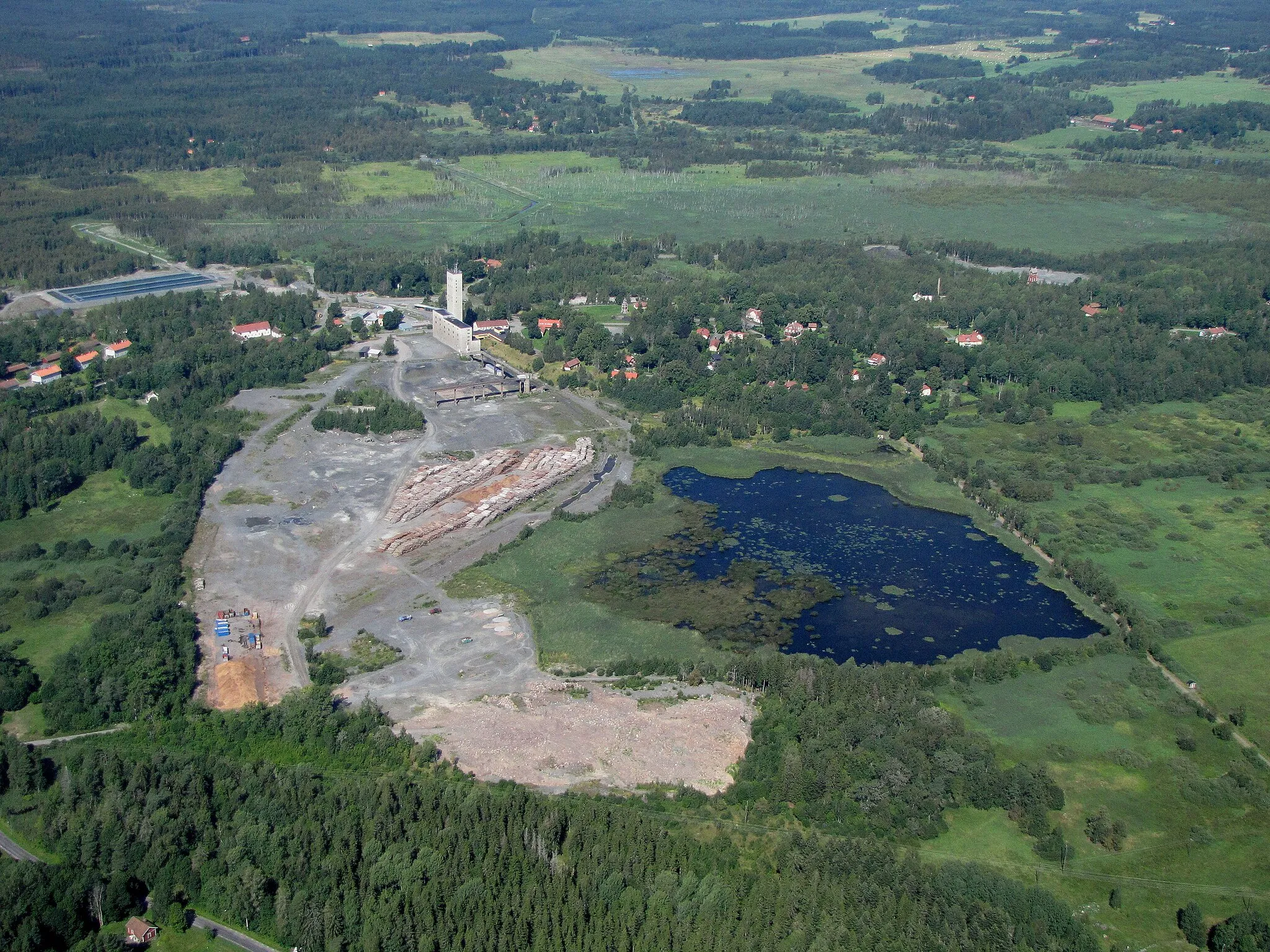 Photo showing: The Dannemora mining area, viewed in NW direction. The historic part is to the right of the modern concrete plant with its high tower. The old wooden bell-tower is seen on the green grass, and the light grey strip behind it is the opencast "Storrymningen". At the far right, there is an old red elevator tower among the trees.