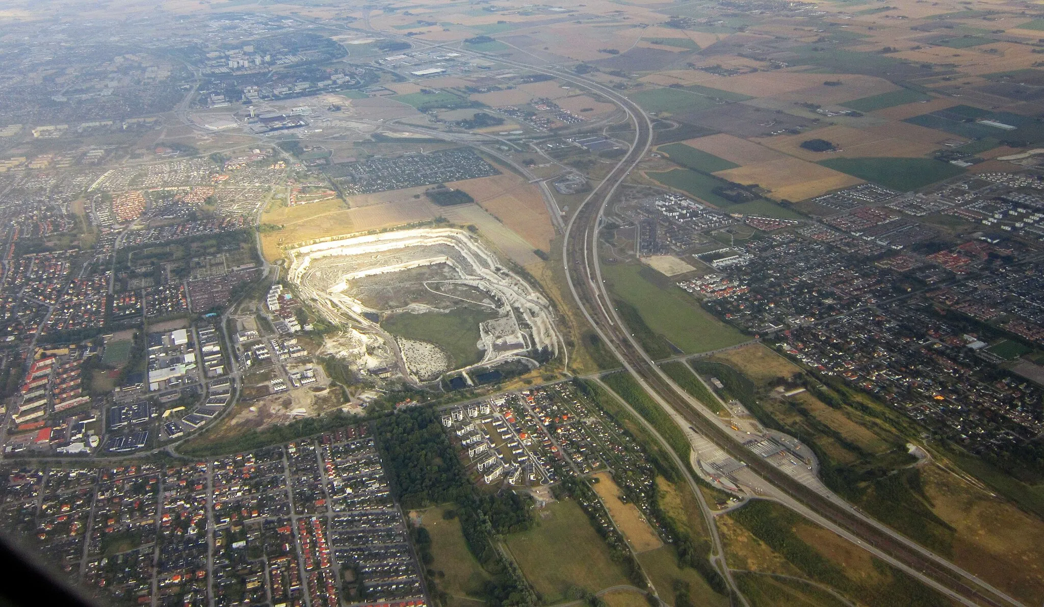 Photo showing: Aerial view of Limhamns kalkbrott. The neighborhood to the left of the motorway (European route E20) is Limhamn, the one to the right is Bunkeflostrand. Toll station for entering Øresund Bridge is visible in the foreground, and Malmö Arena's mushroom-shaped tower -- in the background.