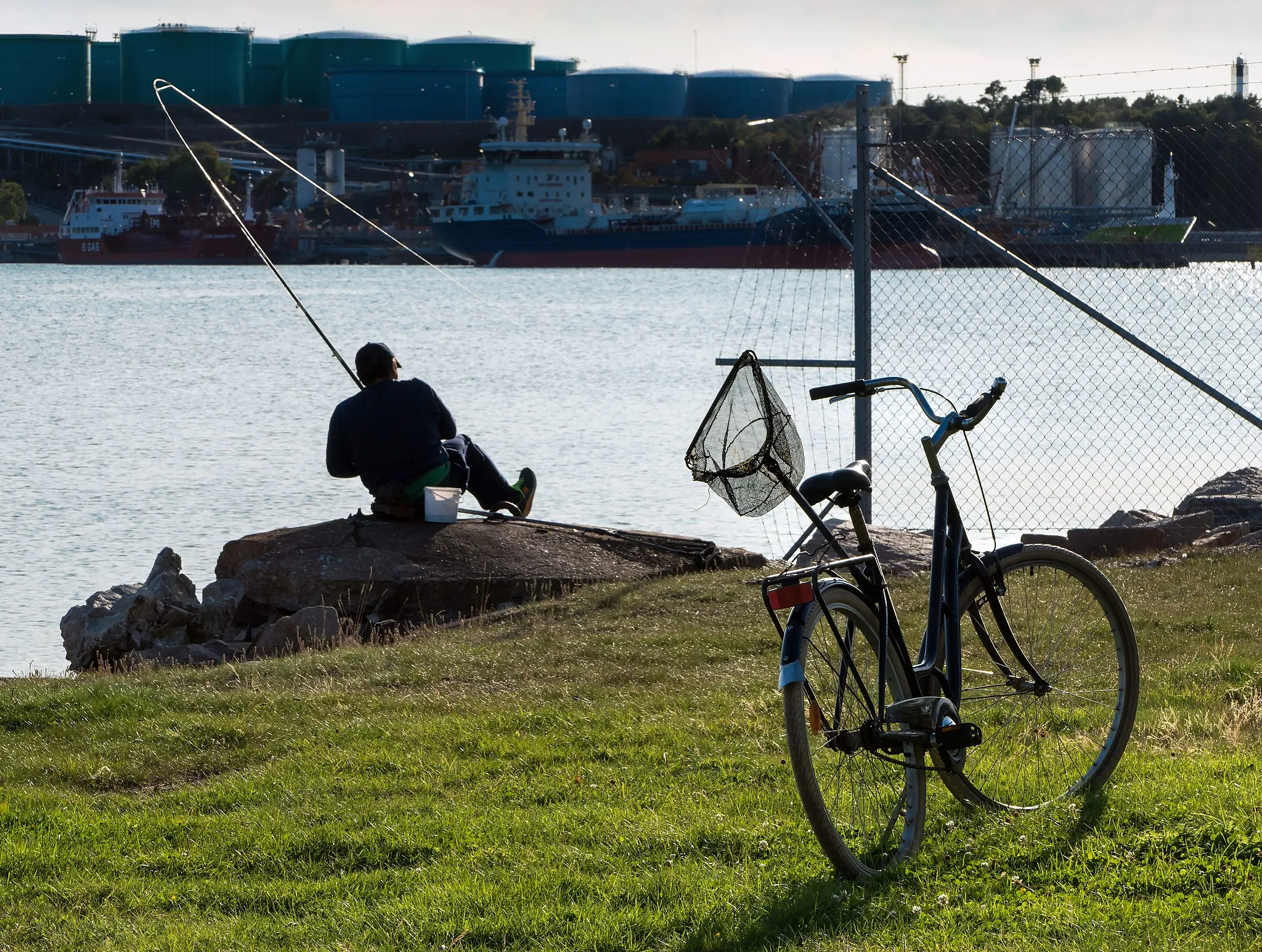 Photo showing: A man fishing in Brofjorden at Lahälla, Lysekil Municipality, Sweden; his bicycle parked next to him. He is just pulling in a fish here. On the oposite shore of the fjord is the Preemraff Oil Refinery.