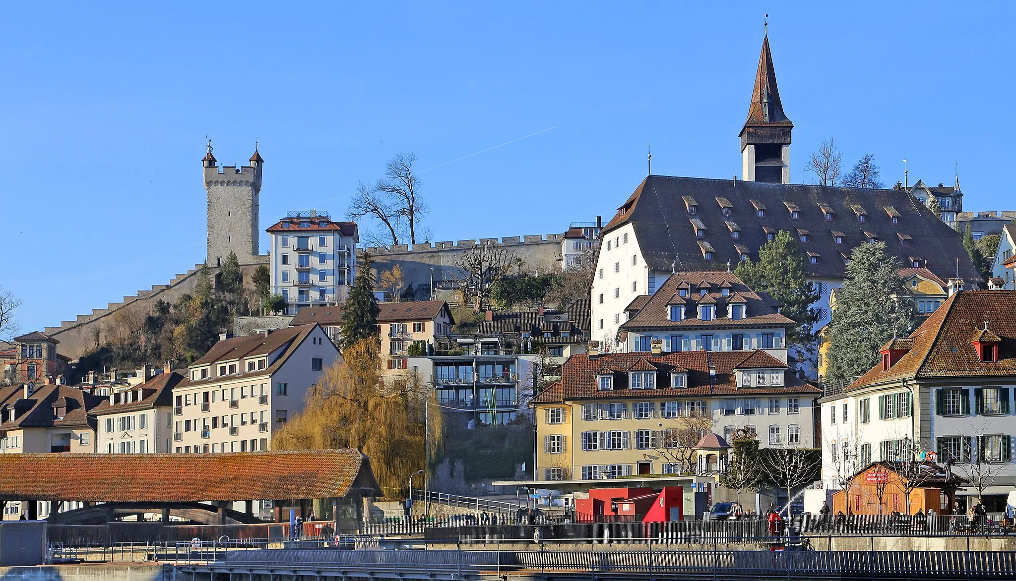 Photo showing: Luzern (Schweiz): Blick auf die Speuerbrücke (gedeckte Holzbrücke) und auf die historische Museggmauer (Stadtmauer) mit dem Männliturm und dem Luegislandturm. Die 870 Meter lange Museggmauer ist ein Teil der ehemaligen Wehrmauer der Stadt Luzern.