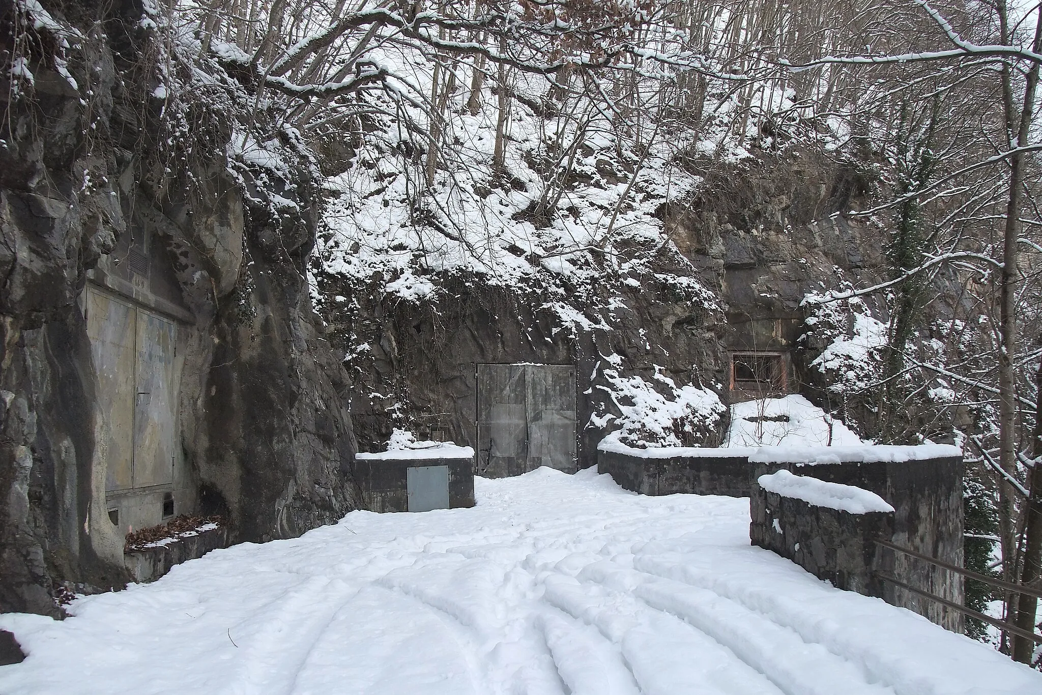 Photo showing: Directly before the entrance a mountain brook flows into the valley. Right of the camouflaged door there are two bunker. One bunker is in the rock beside the door and the other was constructed as toehold below the road. The road was built only for the fortress and ends here at the rock. There is also no trail onward to hike. Sargans, Switzerland, Feb 7, 2013.