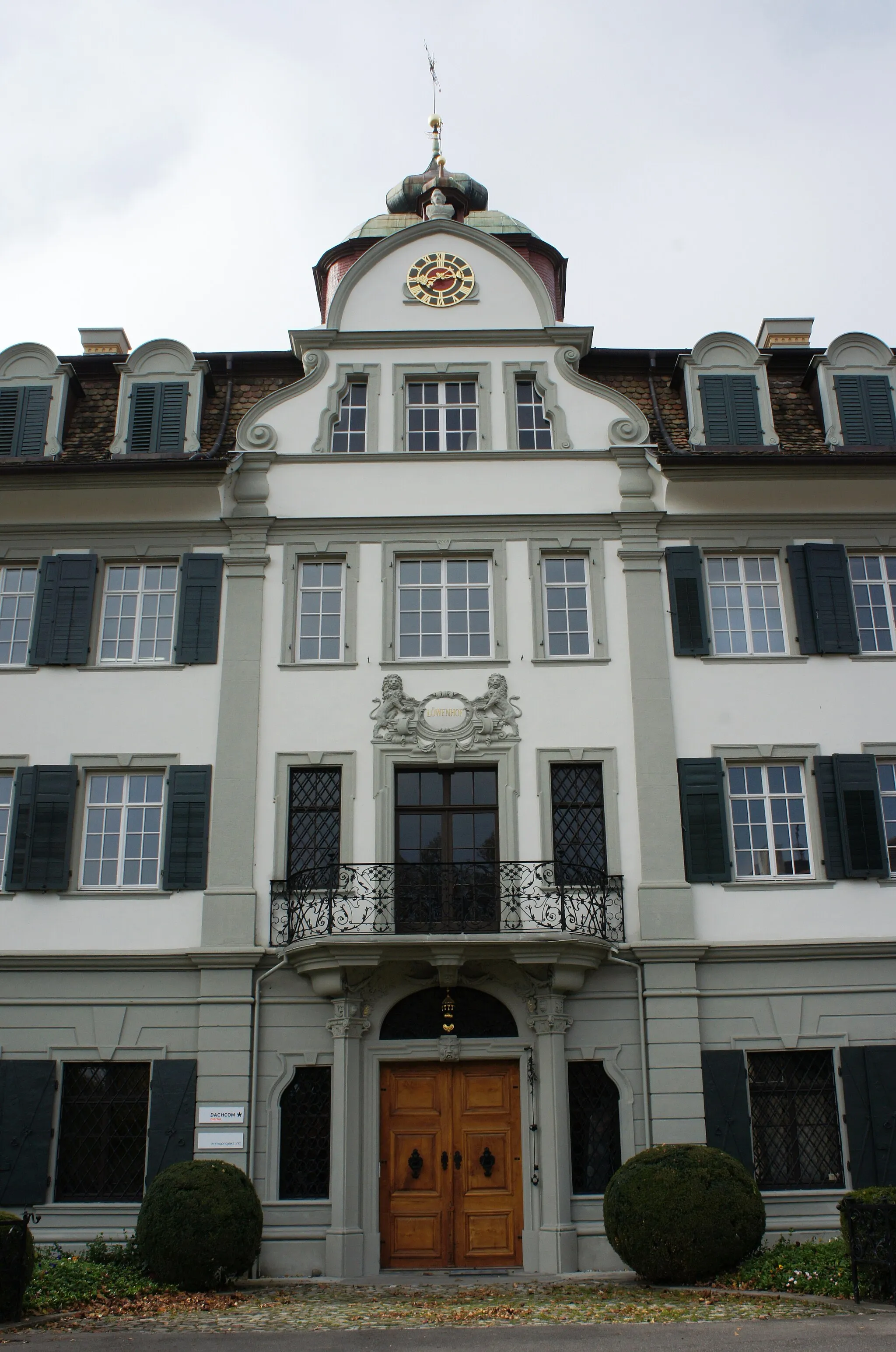 Photo showing: Blick aus Osten auf den Eingangsbereich des Löwenhofes in Rheineck. Das Erdgeschoss ist in grauer Farbe, die beiden oberen Stockwerke haben die Grundfarbe weiss. Die Fensterrahmen und Stuckaturen sind in Grau gehalten. Über der hölzernen Doppeltür ist im ersten Stockwerk ein Balkon mit eisernem Geländer. Über dem vierten (Dach-)Geschoss ist eine Uhr angebracht, vergoldete römische Ziffern und Zeiger auf dunklem Grund, die Zentrumsfläche ist dunkelrot.