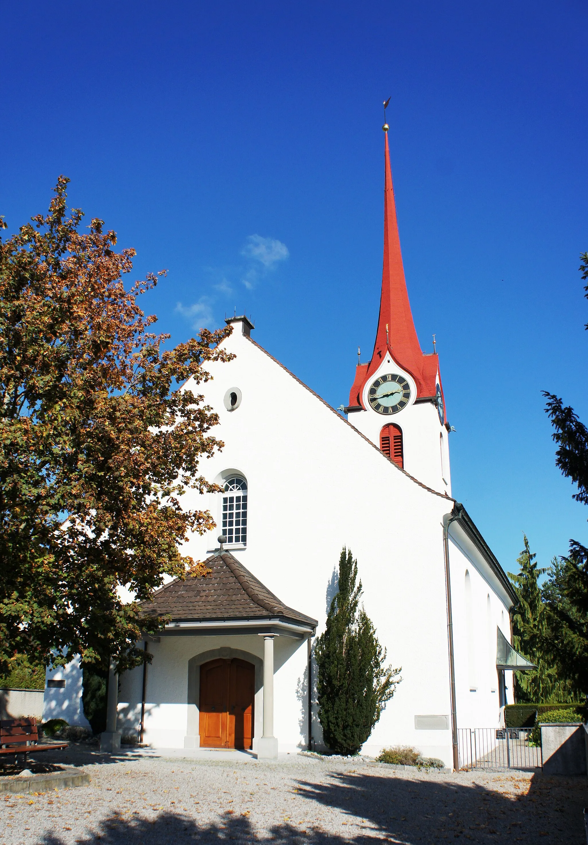 Photo showing: Die ref. Kirche in Rebstein SG aus Südwesten. Im Vordergrund der gepflästerte Vorplatz, in dessen Mitte steht ein Baum mit Sitzbänken umgeben. Stufenlos geht es zum überdachte Haupteingang an der Südwestfassade. Im Zentrum der Nordostfassade steht der Kirchturm mit Uhr und spitzigem, rotem Dach. Zuoberst ist eine Windfahne.