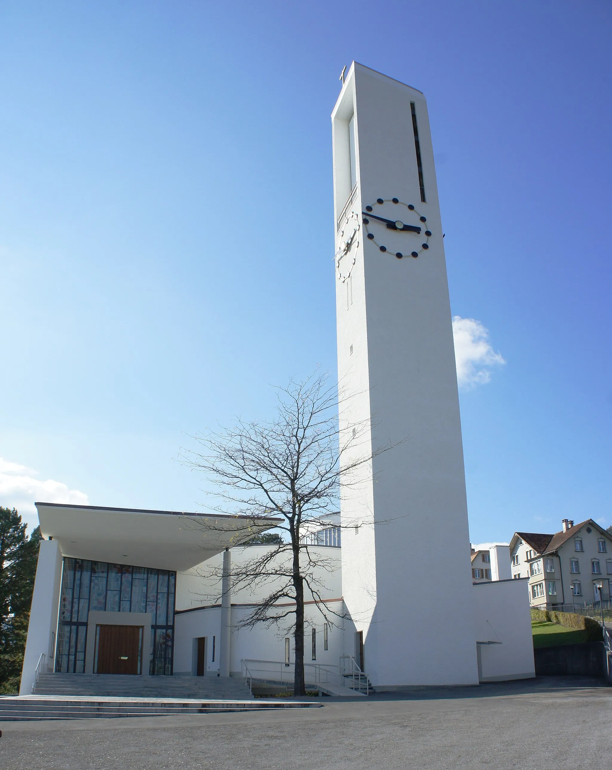 Photo showing: Die kath. Kirche Rebstein aus Osten. Links zum überdachten Haupteingang führen sowohl Stufen als auch eine rollstuhlgängige Rampe. Im mittleren Bildteil steht der Kirchturm mit Turmuhren und Aussichtsplattform auf Höhe des Glockenstuhls. Rechts vom Turm ist die Sakristei. Am rechten Bildrand ist der Burgweg, der zum Friedhof hinauf führt, zu erkennen.