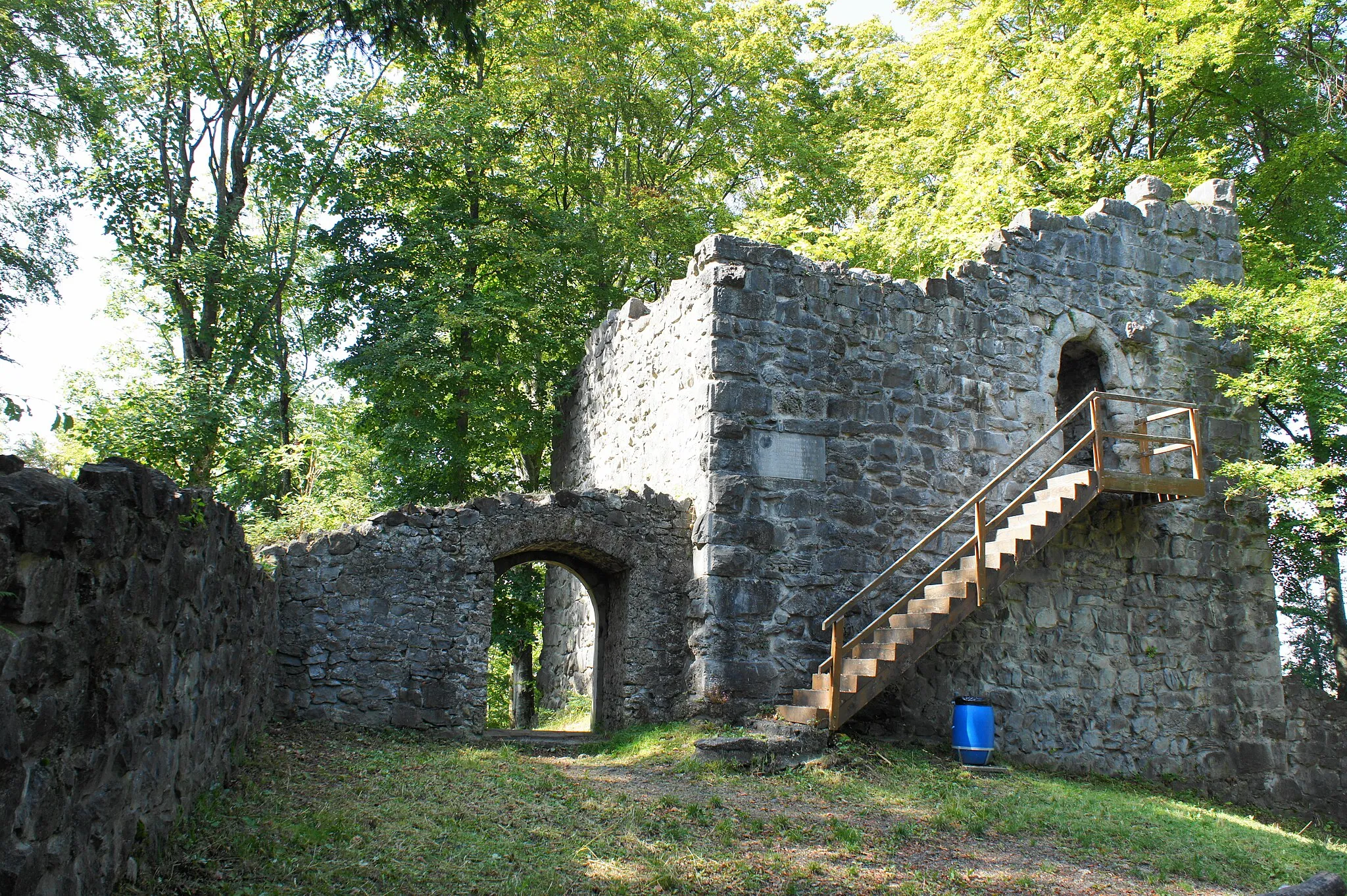 Photo showing: Innenhof der Ruine Ramsenburg, Blick Richtung Eingang (links) und hölzerner Treppe, die zum mit einem Gitter verschlossenen Eingang der Turm-Ruine (rechts) führt, in der Bildmitte eine steinerne Inschrift-Tafel in der Turm-Mauer