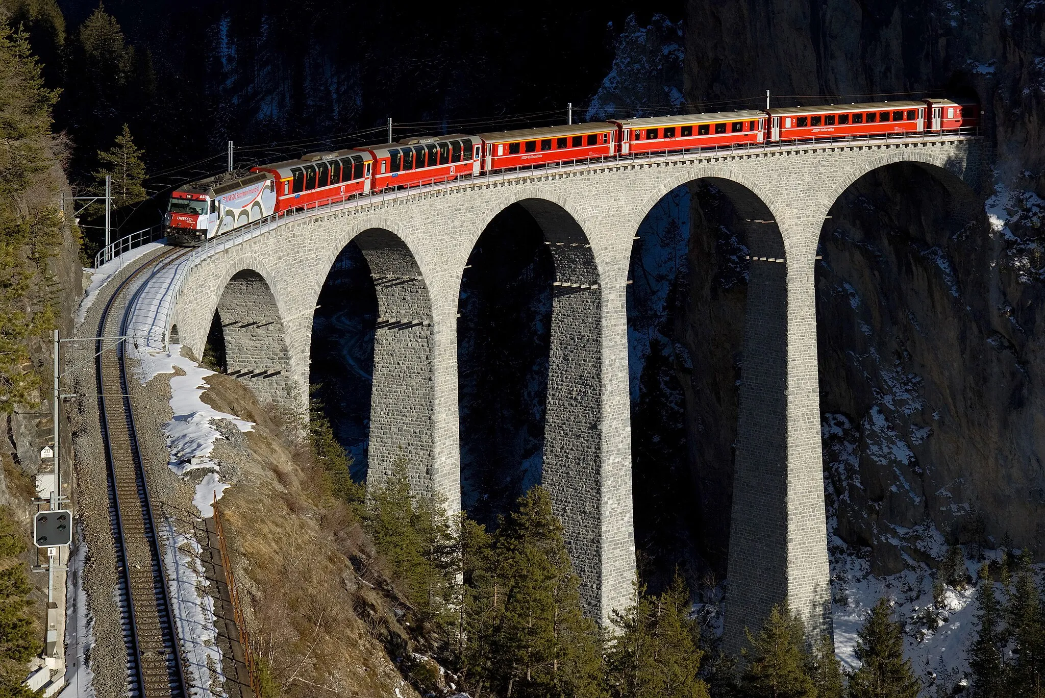 Photo showing: RhB Ge 4/4 III with a Regio Express train from St. Moritz to Chur on the Landwasser viaduct. The locomotive carries advertisement for the UNESCO world heritage Albula and Bernina lines. The stylized viaduct pictured on the locomotive is the very same that it is currently passing over!
Also note the two first panoramic coaches. This type of panoramic coach is normally used on the Bernina line, but because there's less traffic on that line during winter they use the coaches on other parts of the network.