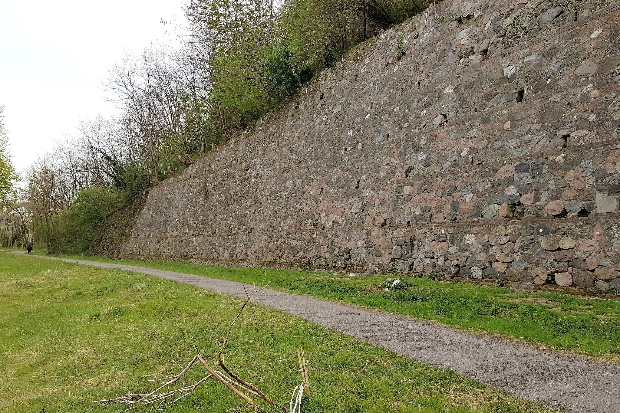 Photo showing: Rock climbing crag made on a containment wall placed next to the river Olona and the ex railway "Ferrovia Valmorea". The wall has spit and top chains and the rock is made of red bricks and porphyry stones. The gravel road in the foreground is the cycleway "pista ciclabile Valle Olona". This wall is located in the municipality of Cairate in the province of Varese in the region Lombardy in northen Italy. Picture taken on the 19th of April in 2021.  2021-04-19