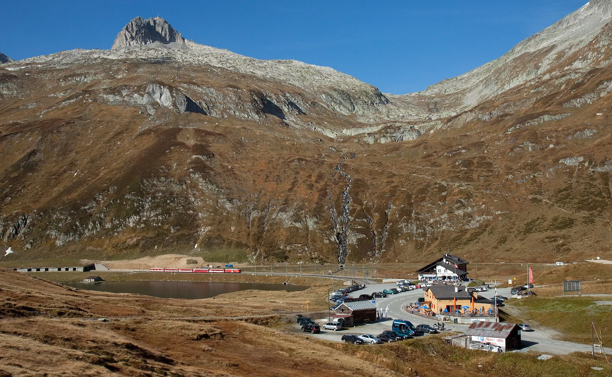 Photo showing: The Oberalp pass summit with the Oberalpsee to the left and some restaurants to the right. A Matterhorn–Gotthard-Bahn local train hauled by a Deh 4/4 motor luggage van of series 21–24 is just arriving at the Oberalppasshöhe station.