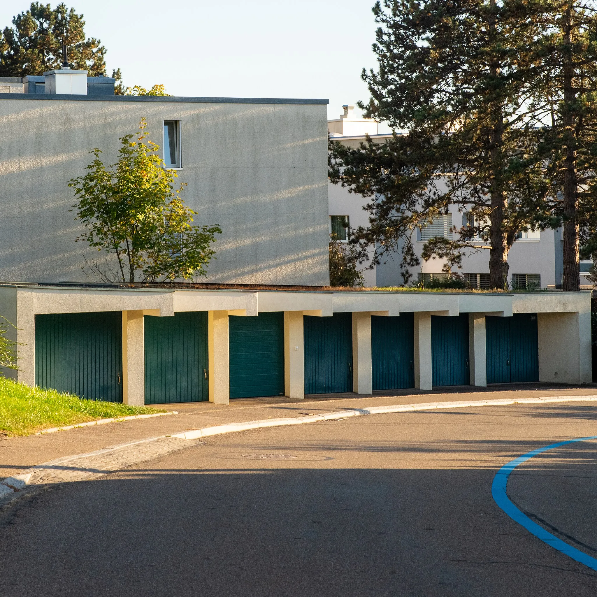 Photo showing: 7 identical garage doors, painted in blue, in the curve of a road with a blue line. There are 3-story residential buildings and pine trees behind the garages.