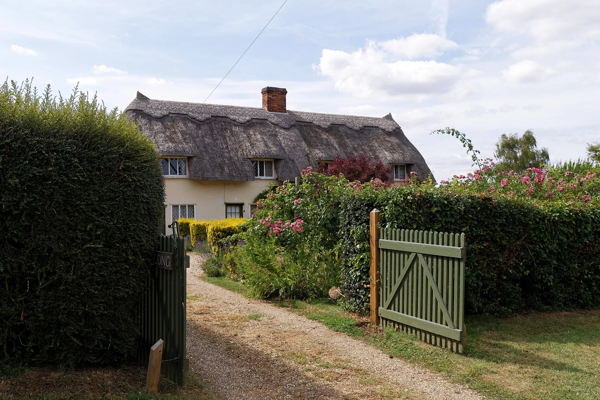 Photo showing: A drive though a gate and hedge, past a shrub garden to a thatched house on Church Street, at Henham, in Essex, England. Software: RAW file lens corrected, optimized and converted to JPEG with DxO OpticsPro 10 Elite, and possibly further optimized and/or cropped and/or spun with Adobe Photoshop CS2.