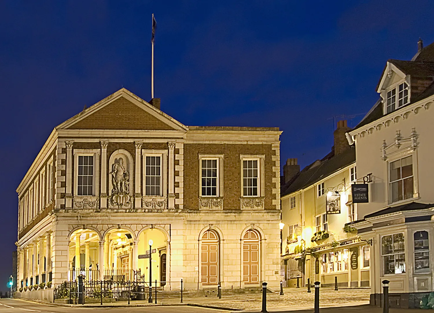 Photo showing: Windsor Guildhall opened in 1689. Night shot showing the  south side with the open ground floor known as the Cornmarket with the statue of Prince George of Denmark above that was installed in 1713. To the right is the 1829 extension. Brown brick and stone.