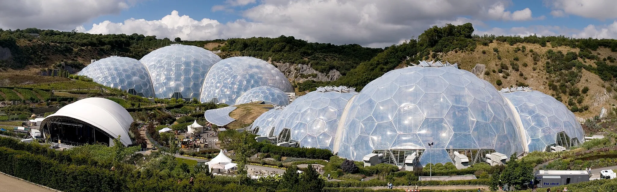 Photo showing: Panoramic view of the geodesic dome structures of the Eden Project. The Eden Project is a large-scale environmental complex near St Austell, Cornwall, England, United Kingdom. The project was conceived by Tim Smit and has quickly become one of the most popular visitor attractions in the United Kingdom. The complex includes two giant, transparent domes made of ETFE cushions, each emulating a natural biome, that house plant species from around the world. The first emulates a tropical environment, the other a warm temperate, mediterranean environment. The project took 2½ years to construct and opened to the public in March 2001.