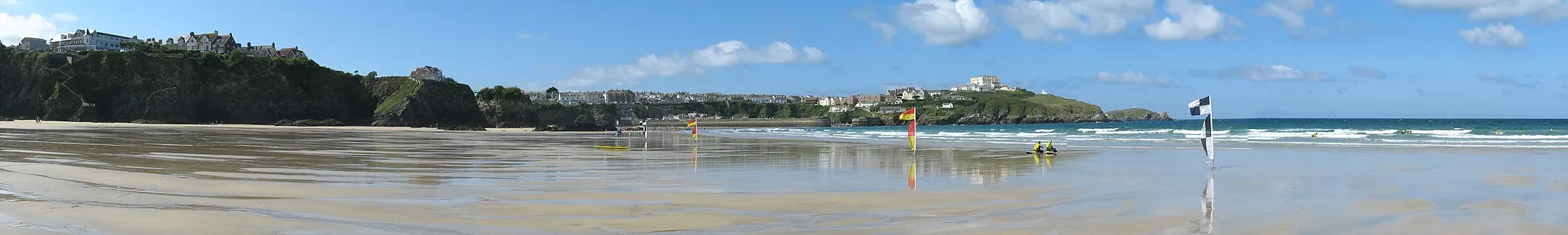 Photo showing: Panorama westward from Tolcarne Beach, Newquay, at close to low-tide. From left to right: Newquay buildings on clifftop; Newquay harbour entrance; The Atlantic Hotel and headland; Towan Head in distance. Two lifeguards sitting on boards in the foreground.