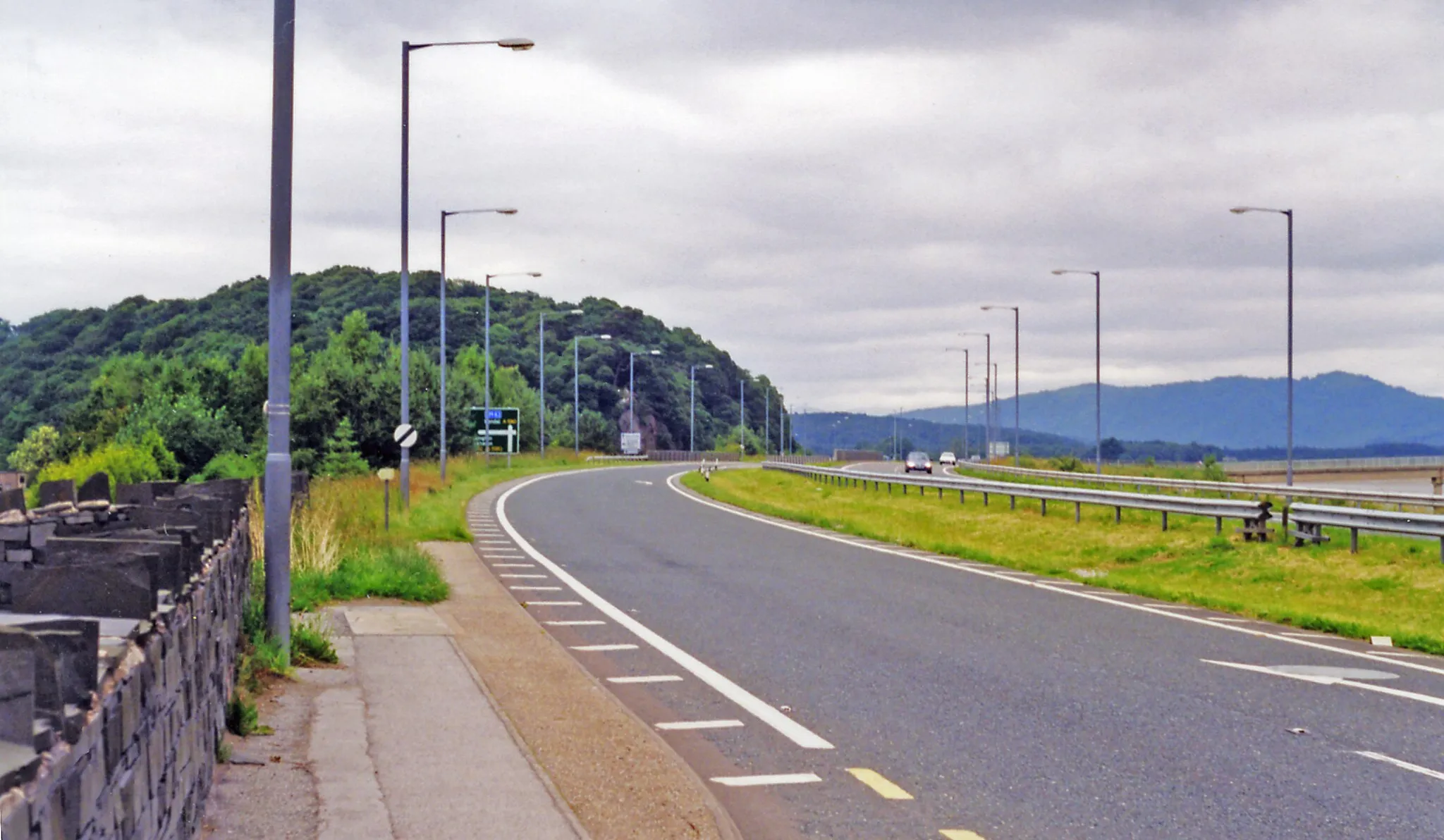 Photo showing: Site of former Greenodd station, on A590, 1998.
View northward, towards Haverthwaite and Windermere Lakeside: ex-Furness Railway, Ulverston - Lakeside branch. On this stretch past Greenodd, the 1970s-widened A590 road has been built along the track-bed, so no trace of the railway seems to remain. Greenodd station closed to passengers from 30/9/46, to goods from 2/12/63. All-year passenger services were run to Lakeside in summer only from 1939, not at all 1941-45, and the line closed completely on 6/9/65. However, from 2/5/73 it has been restored between Haverthwaite and Lakeside by the heritage H&L Railway - which clearly cannot get back through here to Ulverston!