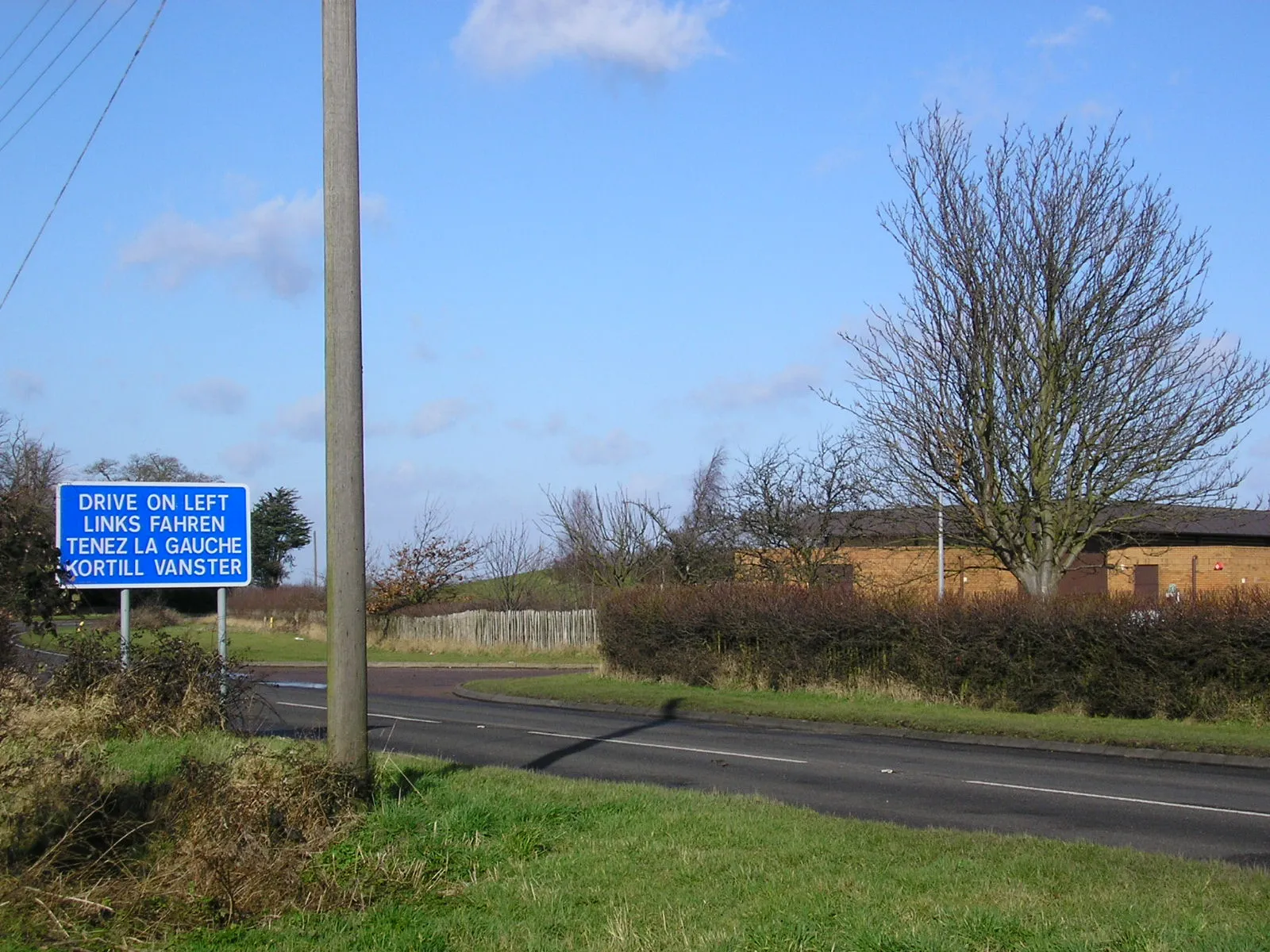 Photo showing: The sign says drive on left in English, French, German and Danish (Körtill vänster). Danish because Harwich has a ferry service to Esbjerg. Clacton Road, Horsley Cross just north of the Wix By-pass (A120). If a foreign visitor having landed at Harwich is at this point they will have just made their first right turn after driving "straight" for 12 km. And possibly there was a serious accident here caused by someone driving on the wrong side. Of course the sign should be on the other side of the road! Essex is not always this helpful - if the foreign visitor continues along this road, they may land up in the Crown Hotel, Manningtree. There, if they go to the toilet, they will have to work out for themselves which of "Gulls" or "Buoys" is appropriate for their sex. The buildings on the right are part of the main treatment works of Tendring Hundred Water Services Ltd - sadly another reference to Tendring Hundred is gone because the company is now called Veolia Water East.