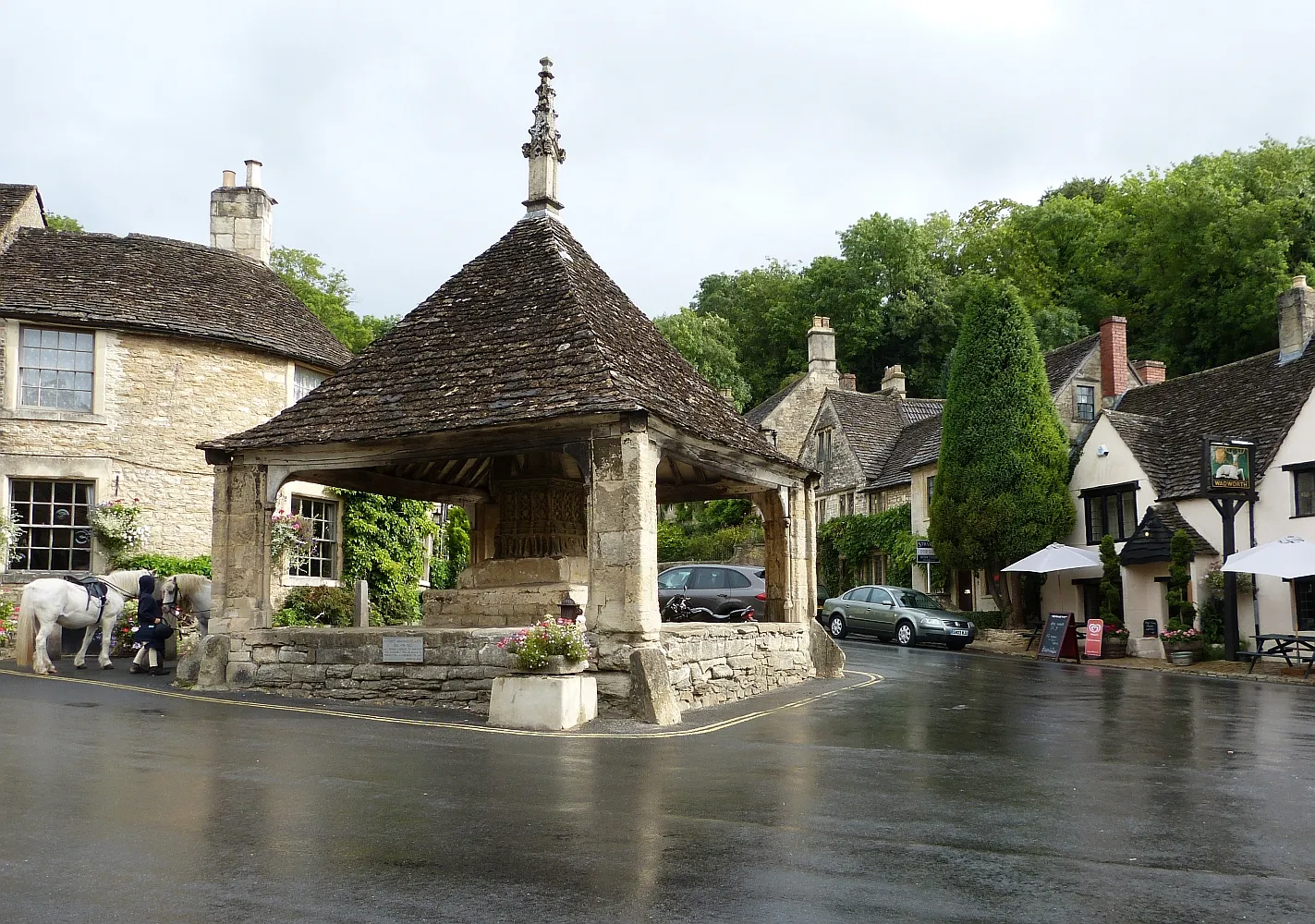 Photo showing: Castle Combe, Butter Cross in The Street. The fourteenth century Market Cross is housed beneath the Butter Cross in the centre of the highly picturesque village of Castle Combe (see shared description). Behind the cross (and behind the ponies) is part of the Castle Inn while the village's other pub, The White Hart can be seen on the right.