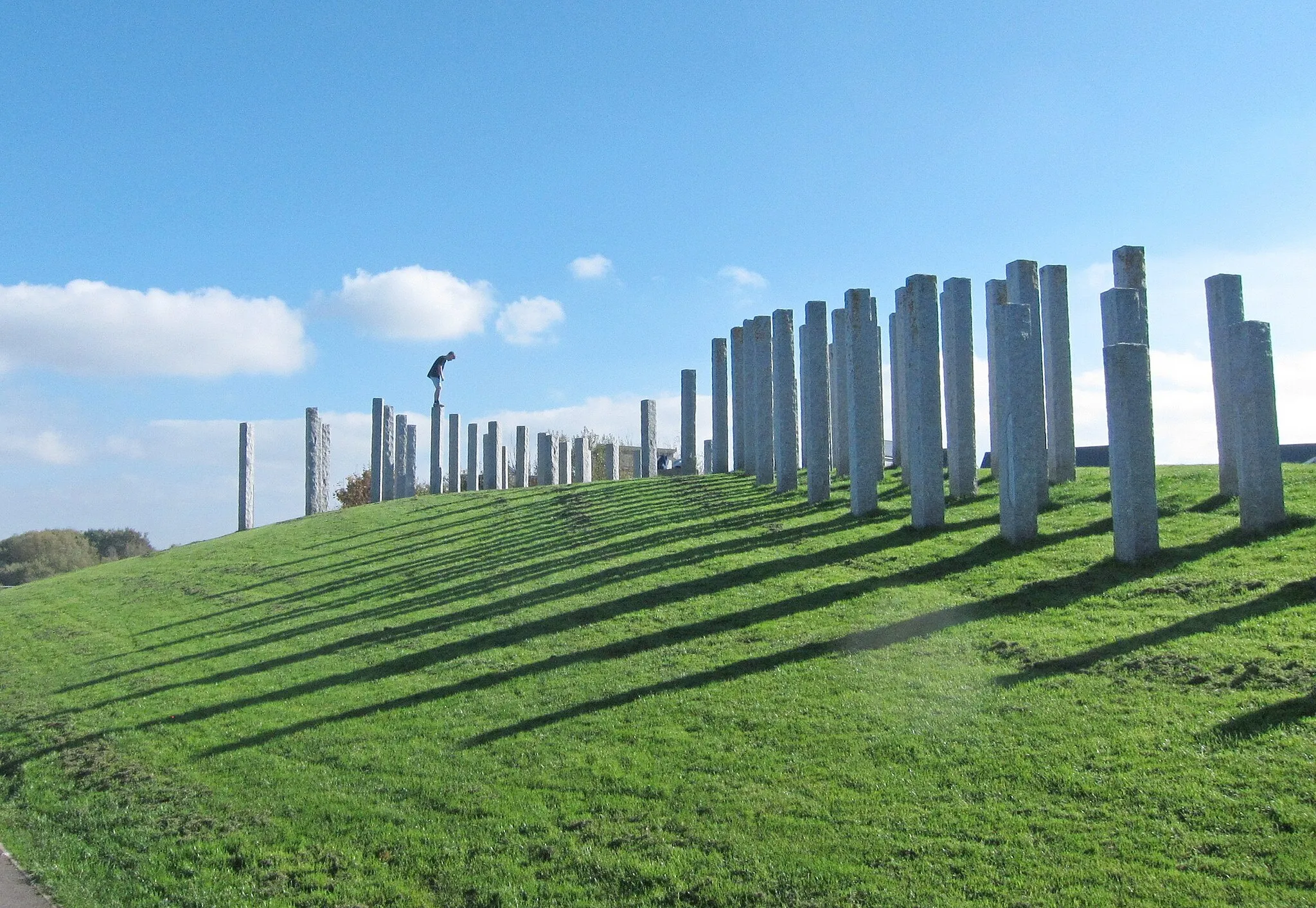 Photo showing: Full Fathom Five sculpture by Michael Dan Archer, at Port Marine, Portishead, near Bristol, Somerset, England. There are 108 granite columns varying in height from 1 to 3 metres arranged across an earth mound, giving views of the Bristol Channel and the coast of Wales.
The pillars were made in a quarry in Xiamen in China and transported by ship to the UK.

The youth is attempting to jump from top to top.