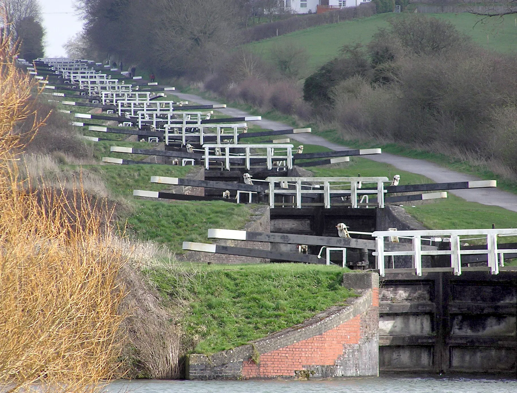 Photo showing: The Caen Hill locks, on the Kennet and Avon Canal, Devizes, Wiltshire, England, in early Spring. The sixteen locks of Caen Hill (seen in this picture), and a further 13 locks, stretch over only 2 miles of the canal. The large side ponds are not seen here. The white "fences" are bridges across each lock.
The building of these locks in 1810 marked completion of the whole 57 mile distance from Newbury to Bath. 
Photographed by Adrian Pingstone in March 2007 and placed in the public domain.
Other versions: