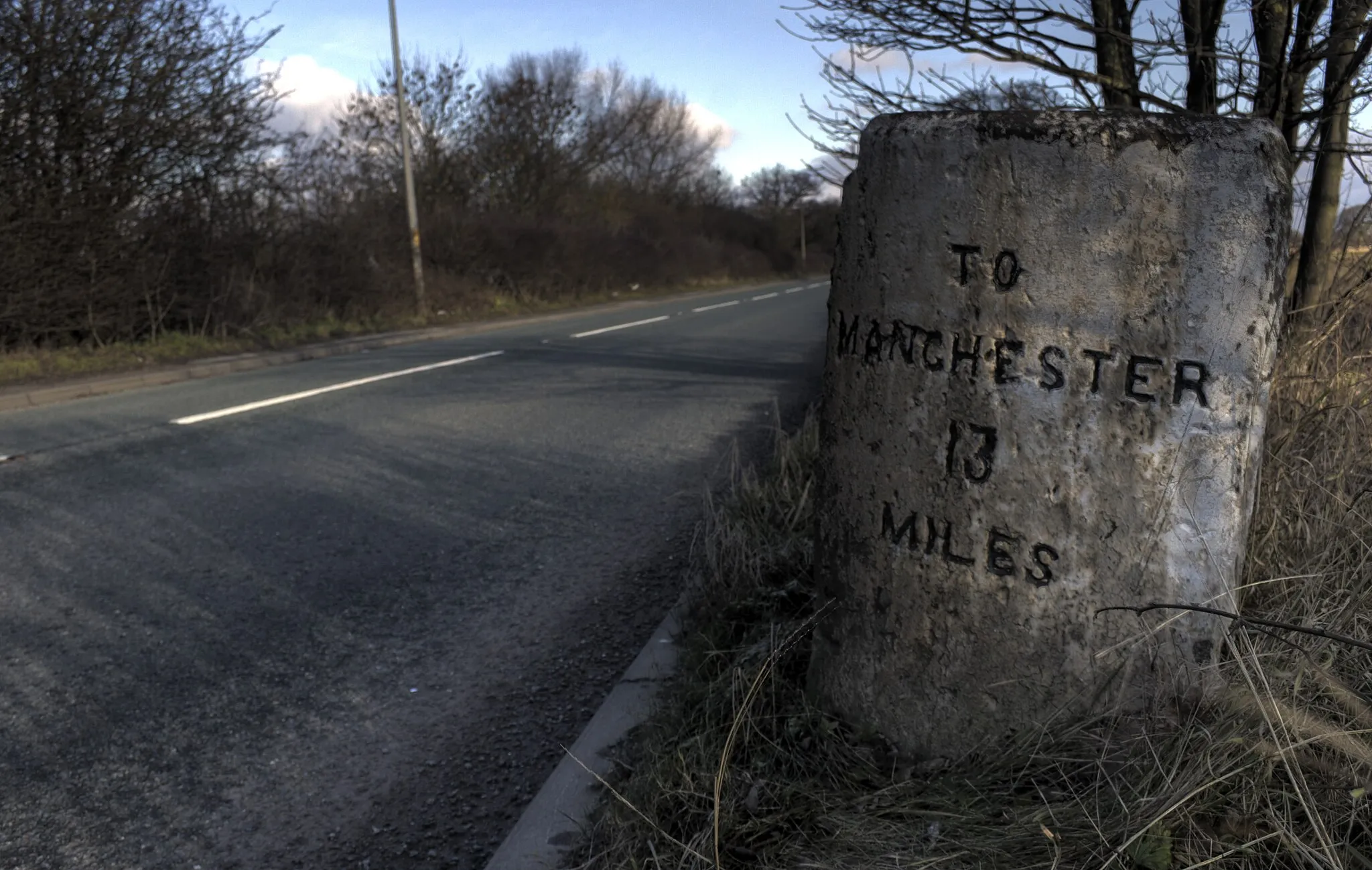 Photo showing: One of a series of milestones on the A57 road near Rixton in Cheshire, between Warrington in Cheshire, and Irlam in Greater Manchester. From the style of lettering, possibly an early 19th century stone probably erected by the Warrington and Lower Irlam Turnpike Trust (this trust was also called the Manchester Road Trust and was only responsible for about 8 miles of road). By the 1820s, many trusts were using cast iron mileposts so it likely dates from around 1810.