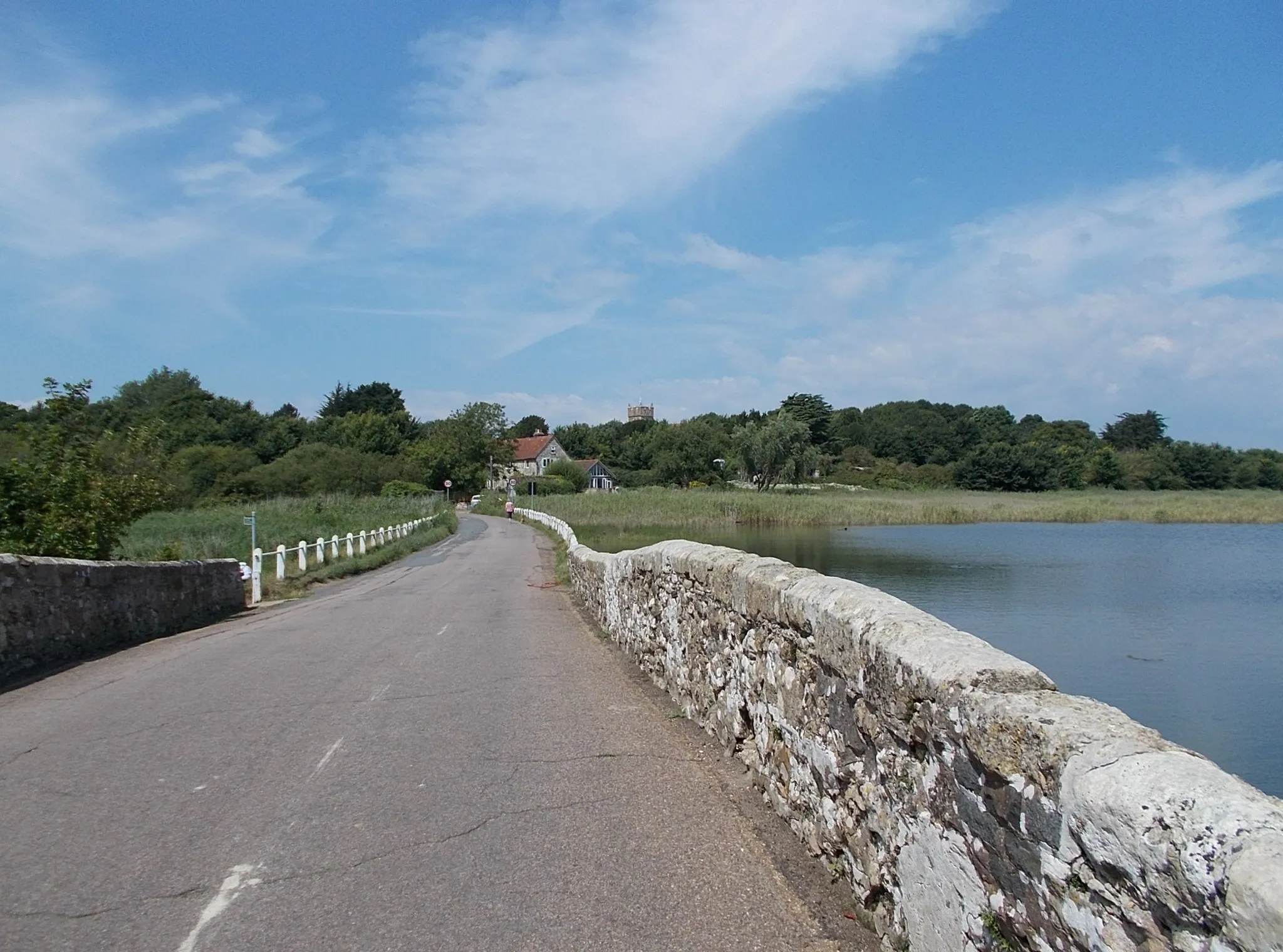 Photo showing: The causeway over the River Yar, leading into Freshwater, Isle of Wight, UK. Looking west, with the tower of All Saints' Church, Freshwater, visible above the trees.