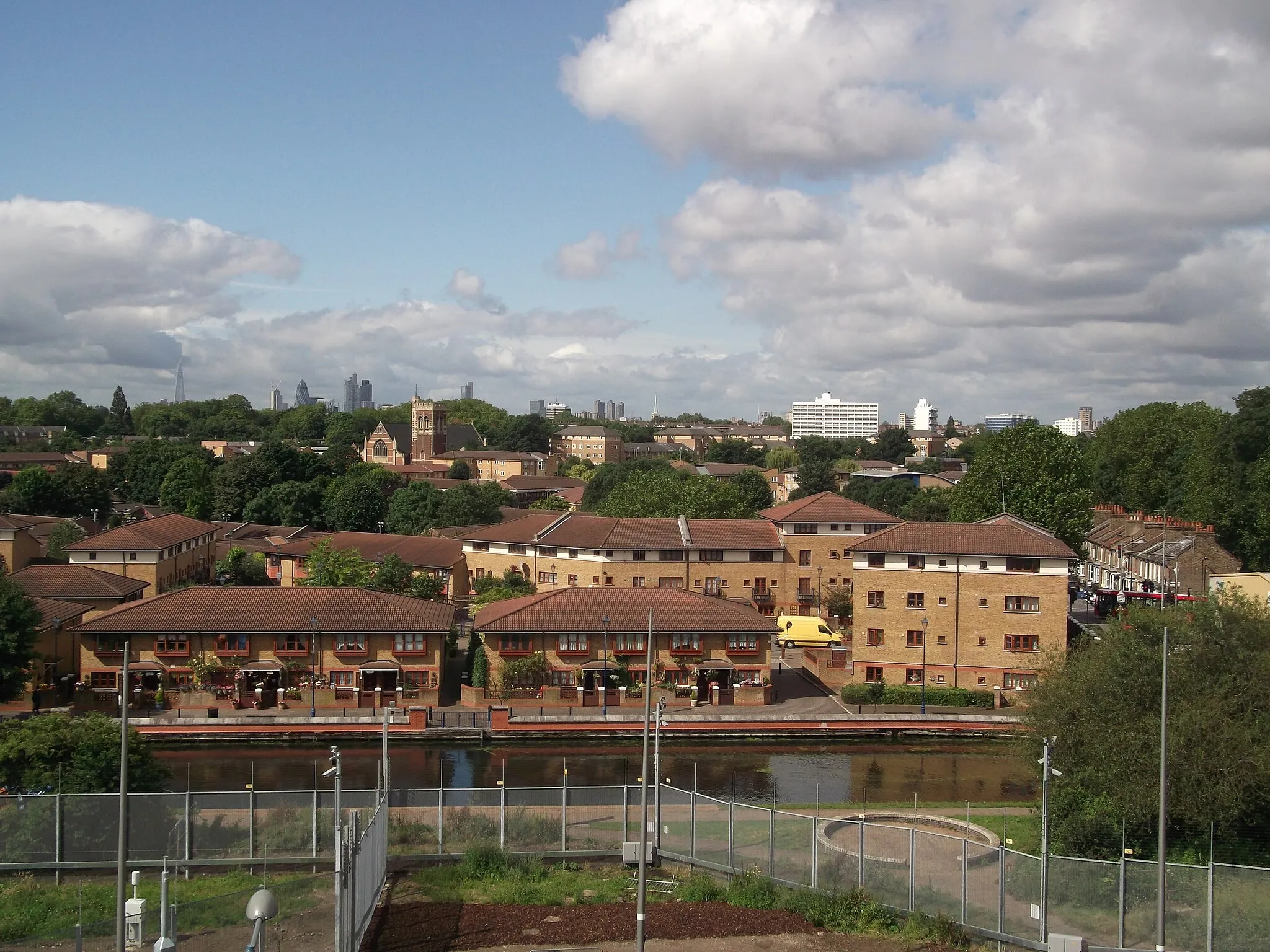 Photo showing: Housing in Hackney Wick. Foreground from left to right: Silk Mills Square; Moat House and Meadow Close. In the distance the skyscrapers in the City of London can be seen, in front of which the tower and gable wall of the church (distinctive red and yellow) belong to St Mary's of Eton, Hackney Wick which won the RIBA architecture award, 2015.