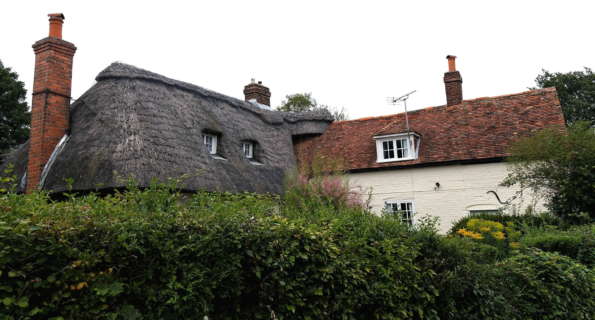 Photo showing: Thatched and tile-roof houses behind a hedge on the lane behind St Nicholas' Church at Barfrestone in Kent, England. Software: RAW file lens-corrected, optimized and converted to JPEG with DxO OpticsPro 10 Elite, and likely further optimized and/or cropped and/or spun with Adobe Photoshop CS2.