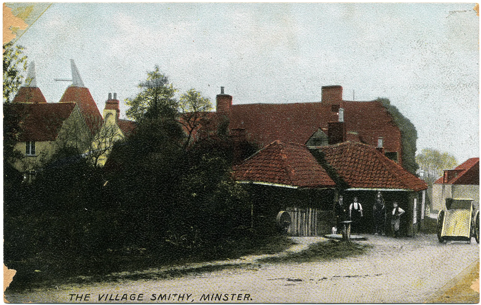 Photo showing: Hand-tinted postcard photo of the smithy (forge) at Minster, Thanet, Kent, England. The photographer was Fred C. Palmer of Tower Studio, Herne Bay, Kent, who is believed to have died 1936-1939. The postcard is believed to be dated 1903-1904 because Palmer moved to Herne Bay in 1903, and because it's printed in Saxony it must be dated before 1914. Also, another card by him in the same style is postmarked 1904. File:Fred C Palmer 008.jpg and File:Fred C Palmer Minster 128.jpg are similarly coloured postcards of Minster by Fred C. Palmer; the three are possibly part of a set. This scene is tidily arranged and posed to show off the smithy's trade. You can just make out the front of a heavy horse, for shoeing, between the two blacksmiths.  The trees have darkened with age, but in front of them, along the side of the roadway, you can just make out the iron machinery and implements waiting for repair. A lot of the work involved fitting the iron tyres round the cartwheels, hence the neat display of a cart on the right.

Border
The remaining border of this image is important for researchers of this photographer. Some photographers trimmed their images more than others, and Palmer has a reputation for producing smaller postcards than other early 20th century UK photographers. He took his own photos, developed them in-house onto postcard-backed photographic paper and trimmed them himself. It is worth adding that during hand-developing the border is actively masked with equipment which both crops the picture and causes the white frame or border to appear on the paper. This frame is part of the design and is one of the reasons why the quality of Palmer's work is so interesting, and why there is an article and category for him on English Wiki. Researchers need to see exactly where the edge of the postcard is, even though in this case there is no white frame.  The edge of the card is the edge of the composition. Thank you for taking the time to read this.