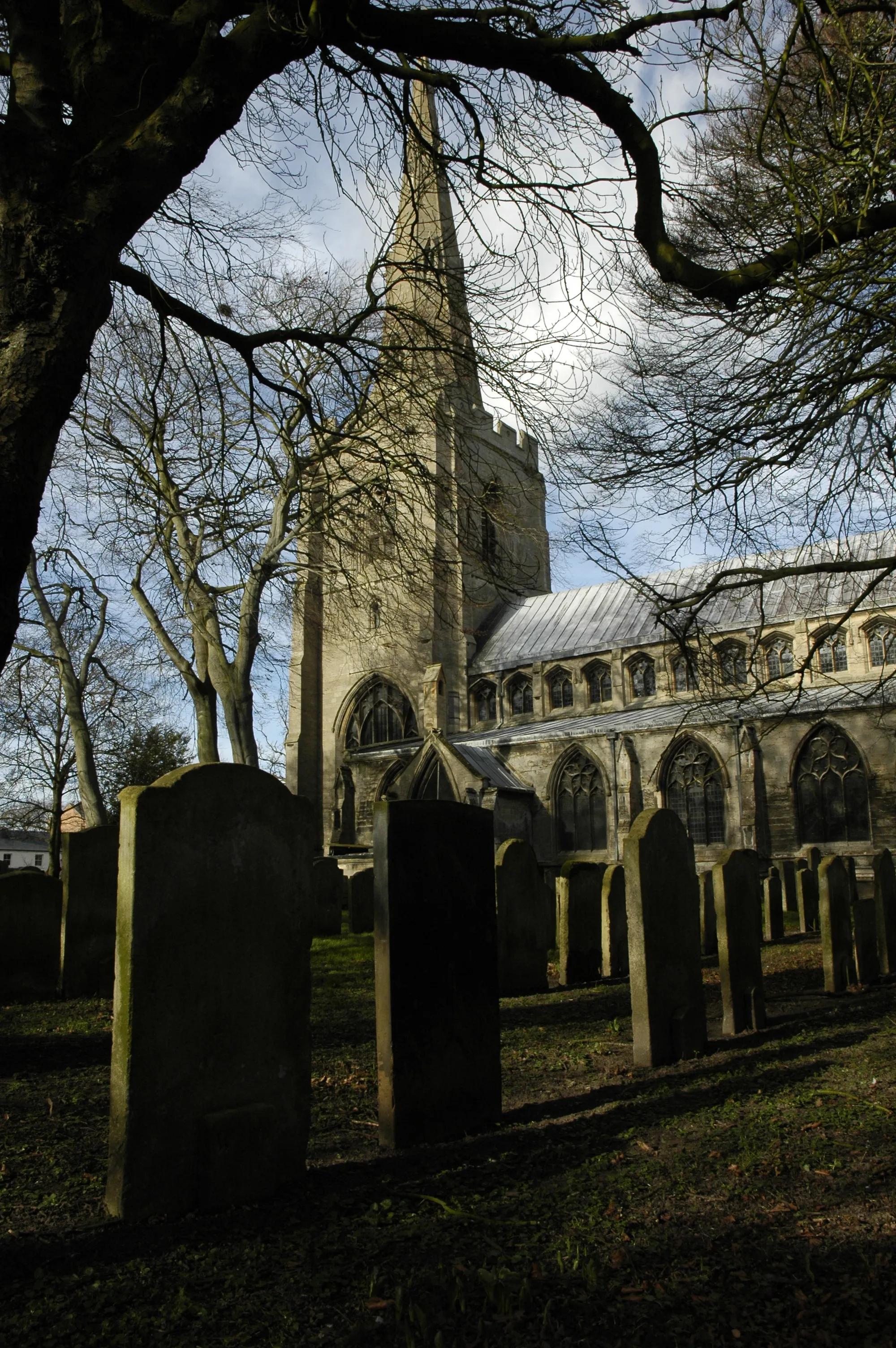 Photo showing: Gravestones in shadow in All Saints' parish churchyard, Holbeach, Lincolnshire, with the west tower and spire in the background