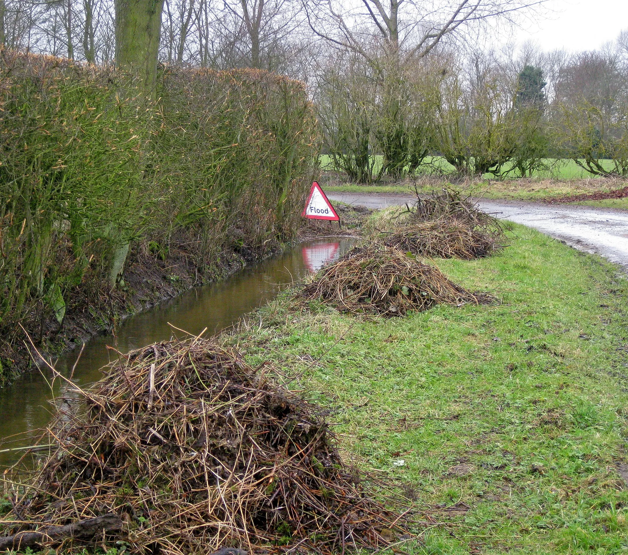 Photo showing: 'February fill dyke'. Hedging and ditching is one of the few jobs which can be done on the farm in February, when the ground is very wet. Of course the wet ends up in the dykes too, which makes the job a little more difficult!The flood sign relates to the road round the corner 1729604