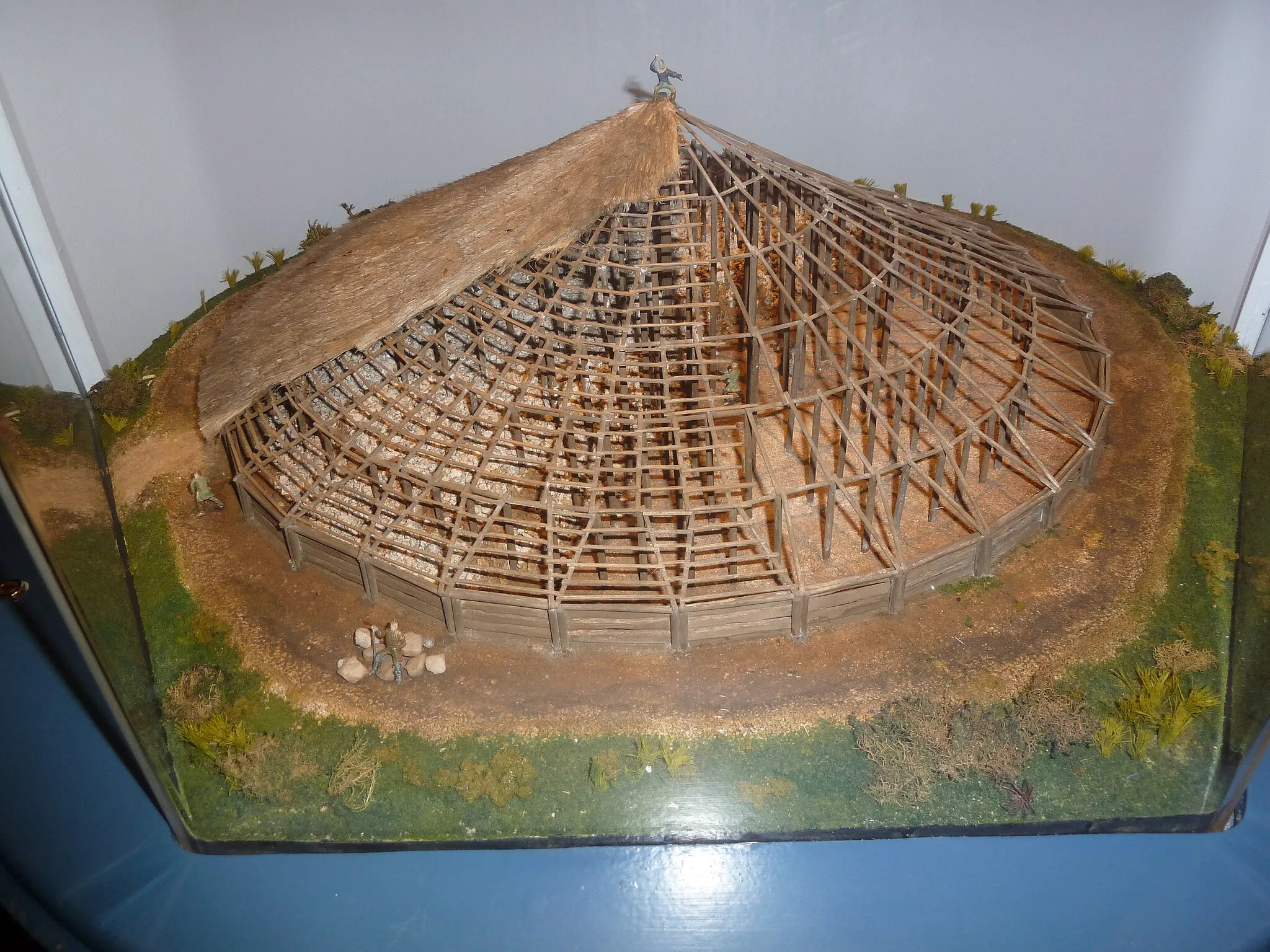 Photo showing: Reconstruction of the Iron age building at Navan, Co. Armagh, Ireland.
Archaeological excavations have revealed that the construction of the 40 metre mound dates to 95 BC (securely dated by dendrochronology).[3] A roundhouse-like structure consisting of four concentric rings of posts around a central oak trunk was built, its entrance facing west (prehistoric houses invariably face east, towards the sunrise). The floor of the building was covered with stones arranged in radial segments, and the whole edifice was deliberately burnt down before being covered in a mound of earth and turf (there is archaeological evidence for similar repeated building and burning of Tara and Dún Ailinne).[3] The bank and ditch that mark the enclosure were made at the same time.