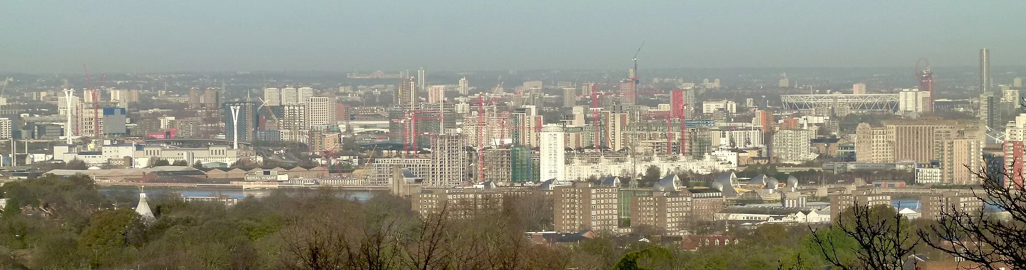 Photo showing: View of the Royal Wharf construction site (Royal Docks, Newham) and parts of East London from Shooters Hill, South East London. In the foreground Woolwich Common, the Rotunda, the Morris Walk Estate and the Thames Barrier. Background right: Stratford.