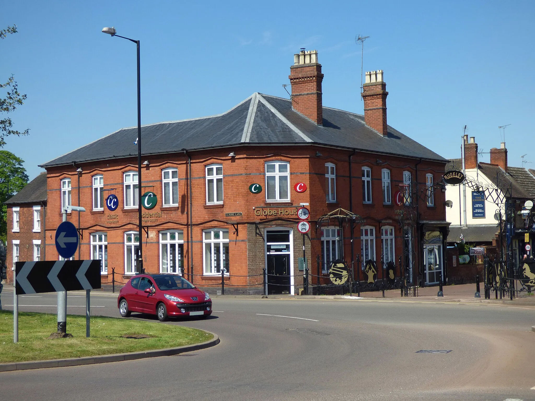 Photo showing: At Globe Island in Rugeley.
The location of the 2015 Miners Memorial sculptures.
The island is at the junctions of Upper Brook Street (pedestrianised), Horse Fair, Sandy Lane, Western Springs Road and Elmore Lane.

Globe House at the corner of Elmore Lane and Upper Brook Street. Estate agents.
Near The Crown pub.
Commemorative Railings

A series of railing posts have been fabricated to resemble nails. Along the main road, these are joined by 'ropes' of wrought iron. Four individual bunches of nails are not part of the railings, and are about half their height. Chains and buckles in wrought iron attach the remaining railing posts to eight egg-shaped medallions suspended midway between each of them. Each of the medallions is decorated with symbols of a different local industry, thereby celebrating Rugeley's heritage. On the left, inwards from the roundabout, they depect mining (pit head wheel, pick, shovel and coal), metal working (knife, forceps and breastplate), and hat-making (a top hat). On the right, they depict a startled horse on its hind legs, glass-making (flasks, vases and a retort), engineering (cog wheel with spikes and a machine tool of some kind), blacksmithing (an anvil, hammer and tongs), and possibly clothing (though the military jacket could alternatively refer to the many army camps in the area). The medallions have the same design on both sides. The designs are almost flat, and are bolted onto the black blackground in each case. The railings with the medallions lead up to a wrought iron archway at the entrance to the pedestrianised area, which has the word RUGELEY in gold in a circular medallion at its apex. There are three horseshoes in a vertical line on each side of the archway. The choice of colour scheme and materials are identical to those for the benches recently installed in Elmore Park nearby, and inscribed with the date '1999'.