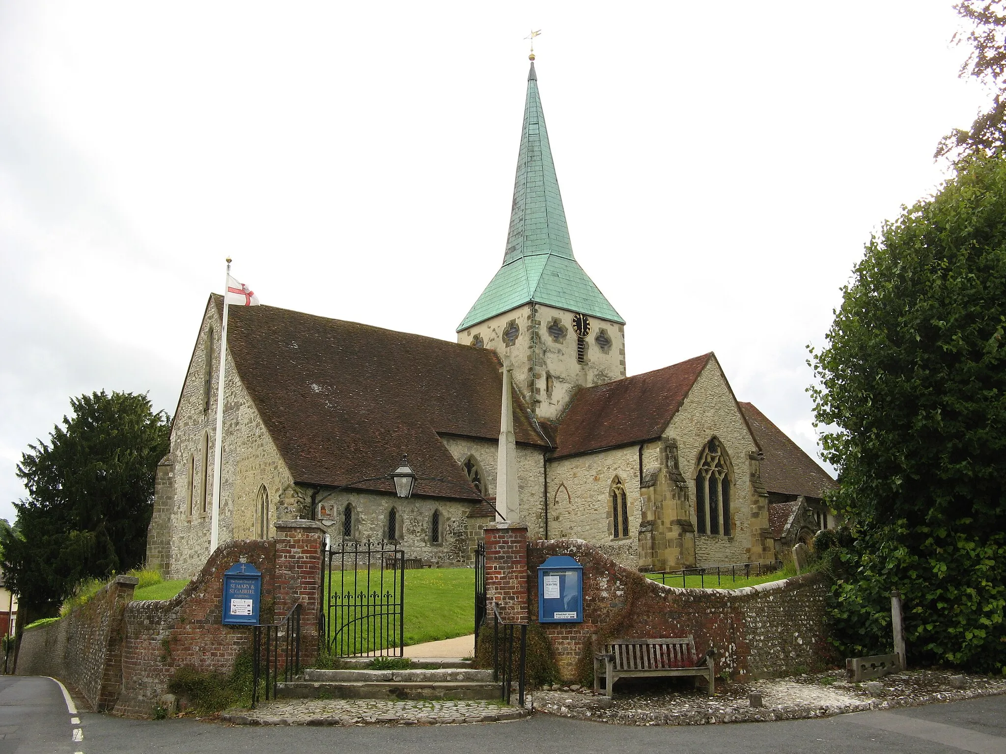 Photo showing: The Parish Church of St Mary & St Gabriel Harting (wording on sign by gate), in the village of South Harting. The tall World War I memorial inside the gate is by Eric Gill.