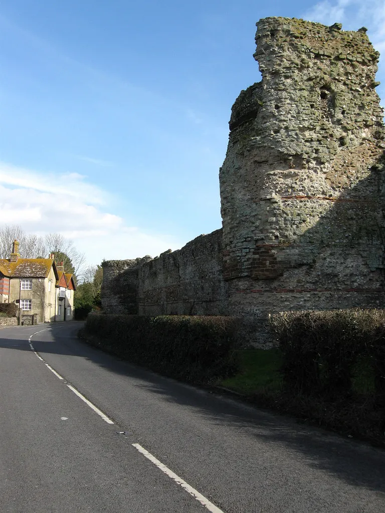 Photo showing: Bastion, Outer Walls, Pevensey Castle Parts of the outer walls date from the 4th century and were built up and strengthened during the medieval period. This bastion was added in the early Tudor period and has modifications from World War Two where the former gaps were filled in and a machine gun post, which is just visible, was added to cover the levels. The house on the left is The Gables and dates from the late 19th century and the road is the B2191 which was the A27 up to 1993.