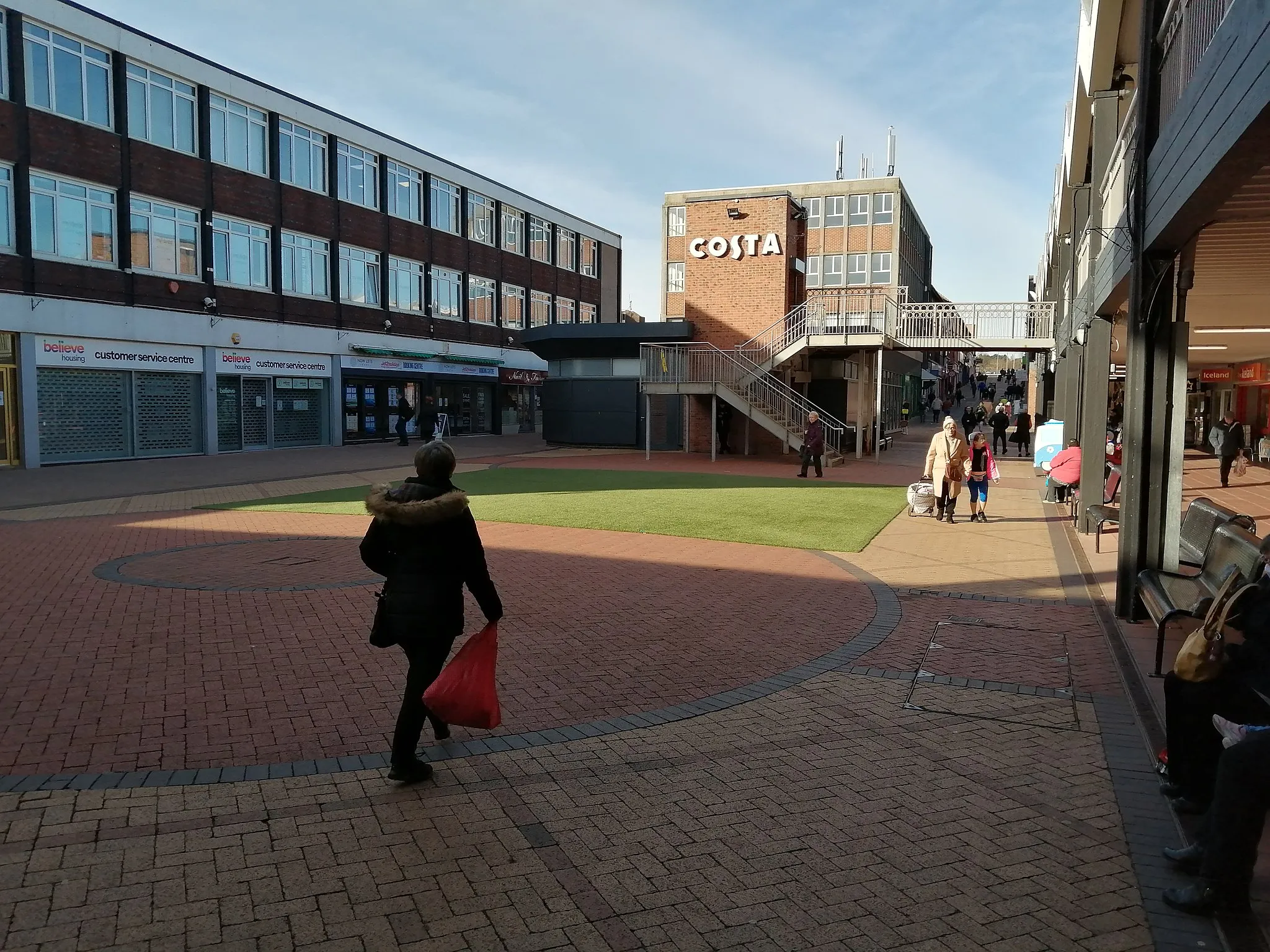 Photo showing: The large pedestrian area at Castle Dene Shopping Centre in Peterlee. The open area is paved with circular patterns in the brickwork, and a large green patch of synthetic turf has been laid down further ahead. Behind this is the building housing a public access lift and stairway, its wall bearing the signage of the Costa Coffee shop behind it.
To the viewer's left is the Believe Housing customer service centre, currently closed as a result of the COVID-19 pandemic, A Jet2Holidays travel centre, and a nail salon called Nail Fairy.

To the right, the signage for an Iceland supermarket and Superdrug can just be seen. Various pedestrians can be seen going about their business. Up ahead, the pedestrianised area extends uphill to more shops.