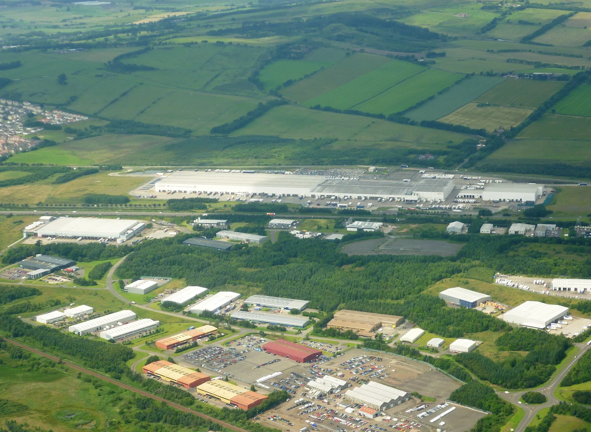 Photo showing: Tesco's Distribution Centre in Livingston (the long white building) opened in 2007, supplying all the company's stores in Scotland and Northern Ireland. It covers an area of one million square feet. The road haulage company Stobart transports goods from the centre to the Grangemouth rail terminal, whence they are sent on by rail to Inverness for distribution throughout the north of Scotland.