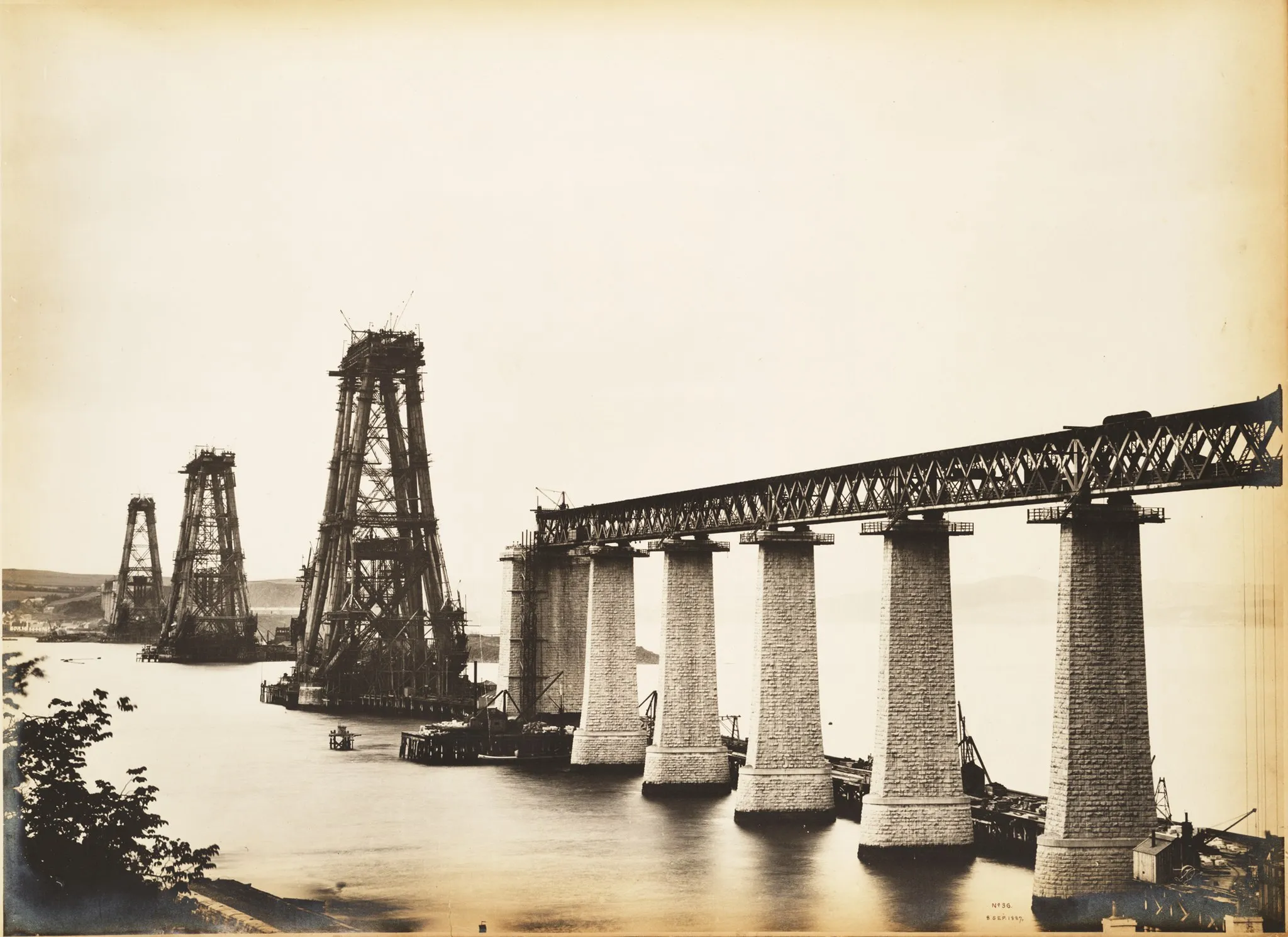 Photo showing: Photograph of the Forth Bridge under construction, general view from back of Newhalls Inn, South Queensferry. A view obtained to meet a continual demand for a picture showing more of the approach viaduct and less of the bridge itself. To this end it was necessary to take up a position which would considerably foreshorten the whole structure. The three cantilevers are shown quite clear of each other, and the left of the picture is bounded by a portion of the Hawes Pier. The detail of viaduct piers can here be seen to perfection, including the platforms used in connection with lifting arrangements. The piers in the foreground have a measurement of rather more than 2 inches at the base, and are about 7 inches in height, the girder about 1.1/5 inches, the Queensferry cantilever 3 inches at the base and 6.1/5 inches high. With these dimensions there should be no difficulty in forming a correct idea of the scale of this really excellent photograph. Transcription from: Philip Phillips, 'The Forth Railway Bridge', Edinburgh, 1890.