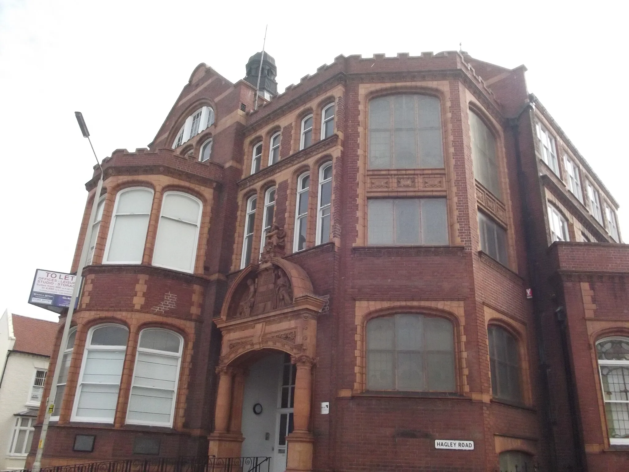 Photo showing: Victorian buildings at the bottom of Hagley Road in Stourbridge, West Midlands.
This building is the former Free Library & Technical College.
On the corner of Church Street and Hagley Road.
It was Grade II listed in 1989 under the title Stourbridge College of Art.
But the whole building is currently To Let, for office, leisure, studio and storage usage.
Stourbridge College of Art, Dudley
HAGLEY ROAD
1.
5106
SO 9084 1/103 Stourbridge College
of Art
GV
II
2.
College of Art, formerly town library and technical college. 1903-4 and 1908-9 by
Frederick Woodward. Red brick with terracotta dressings and Cumberland and Welsh slate
roofs with various brick stacks. In Netherlandish Renaissance style. Complex plan
with various ranges occupying corner site. Mostly of 3 storeys, basement and attic.
Entrance to corner on right has elaborate terracotta archway surmounted by relief figures
within a tympanum. Steps lead to part-glazed doors filled with elaborate stained glass
including portrait roundels of Kelvin, Shakespeare, Rubens and Mozart. Tall basket-
arched windows over, with moulded terracotta frames and ornamental cornice hoods.
Coped Dutch gable. To right a canted battlemented section containing the staircase
with similar elaborately ornamented window frames. Further to right, facing Hagley
Road, a 2-storey range in similar style. On right an elaborate terracotta archway
leading to door, and a clock tower over with clock faces within balconied arches and a
domed open belvedere on top. Behind this section rises the second span of the range facing
Church Street. Wide sash windows, some with terracotta decoration. On the roof ridge
an elaborate domed lantern. To left of the front entrance the end of the range facing
Church Street which has a large polygonal 2-storey bay with battlemented parapet and
large arched window over. To left the front facing Church Street which is a 9-window
range with 4 slightly projecting gabled sections and similar fenestration with elaborate
terracotta window surrounds. Further doorway in 2nd gable from left. 2 elaborate
domed lanterns on roof. Interior: glazed doors and partitions, staircase with
decorative cast-iron balustrade and a fine and extensive series of stained glass windows
in art-nouveau style, one signed S Evans, Stained Glass Works, West Smethwick. These
are most elaborate on the stairs but continue on both the Hagley Road and Church Street
fronts and are a very important feature of the building. Andrew Carnegie contributed
£3,000 to the library's foundation and a further £700 to the 1908 newsroom extension.

Listing NGR: SO9038184050