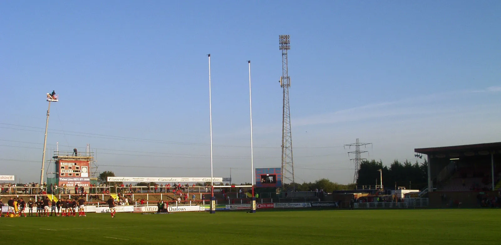 Photo showing: Stradey Park, Llanelli prior to the Magners League rugby match between Llanelli Scarlets and Glasgow Warriors. The image shows the East Terrace. On the top of the cherry picker towards the left of the photo is a BBC cameraman. To the right is the scoreboard. On the pitch below these, the Scarlets team are warming up. Note the 'sosbans' (saucepans) on top of the goalposts. The blue box to the right of the goalposts is the television studio. The outside broadcast trucks can be seen below the electricity pylon.