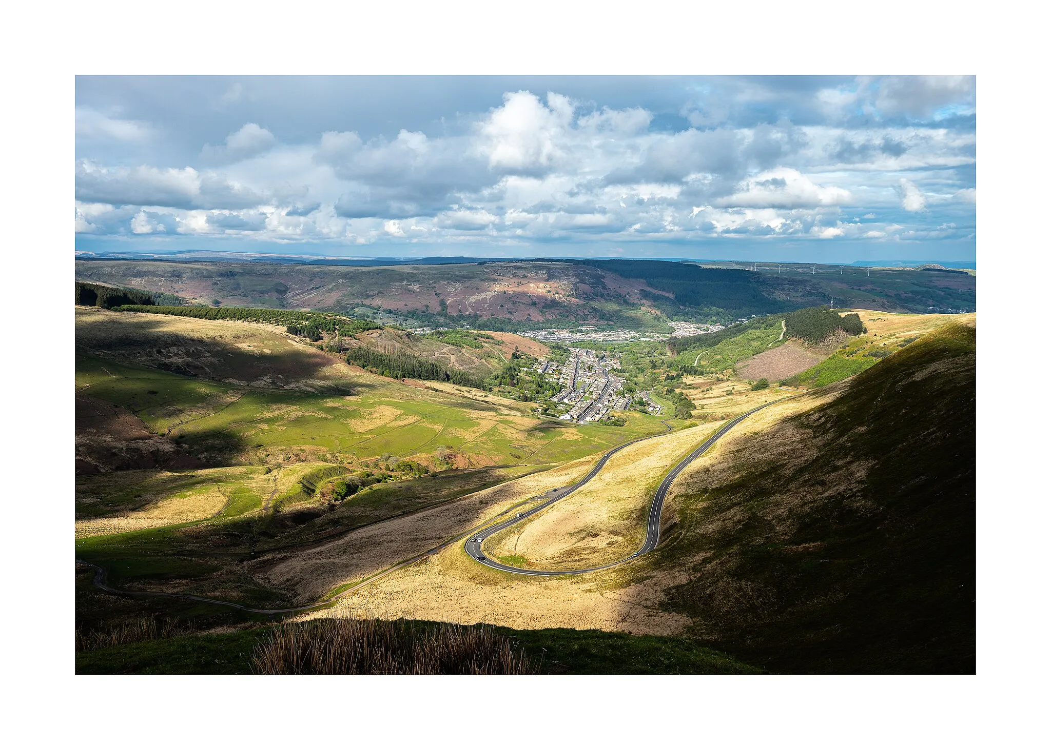 Photo showing: Cwmparc. The view from the Bwlch mountain overlooking the former coal mining community of Cwmparc, Treochy in the Rhondda Valley. In the distance is the Brecon Beacons National Park. This photo was taken during a brief break in the cloud which illuminated the valley and mountain road, with some cloud shadows present in the foreground.